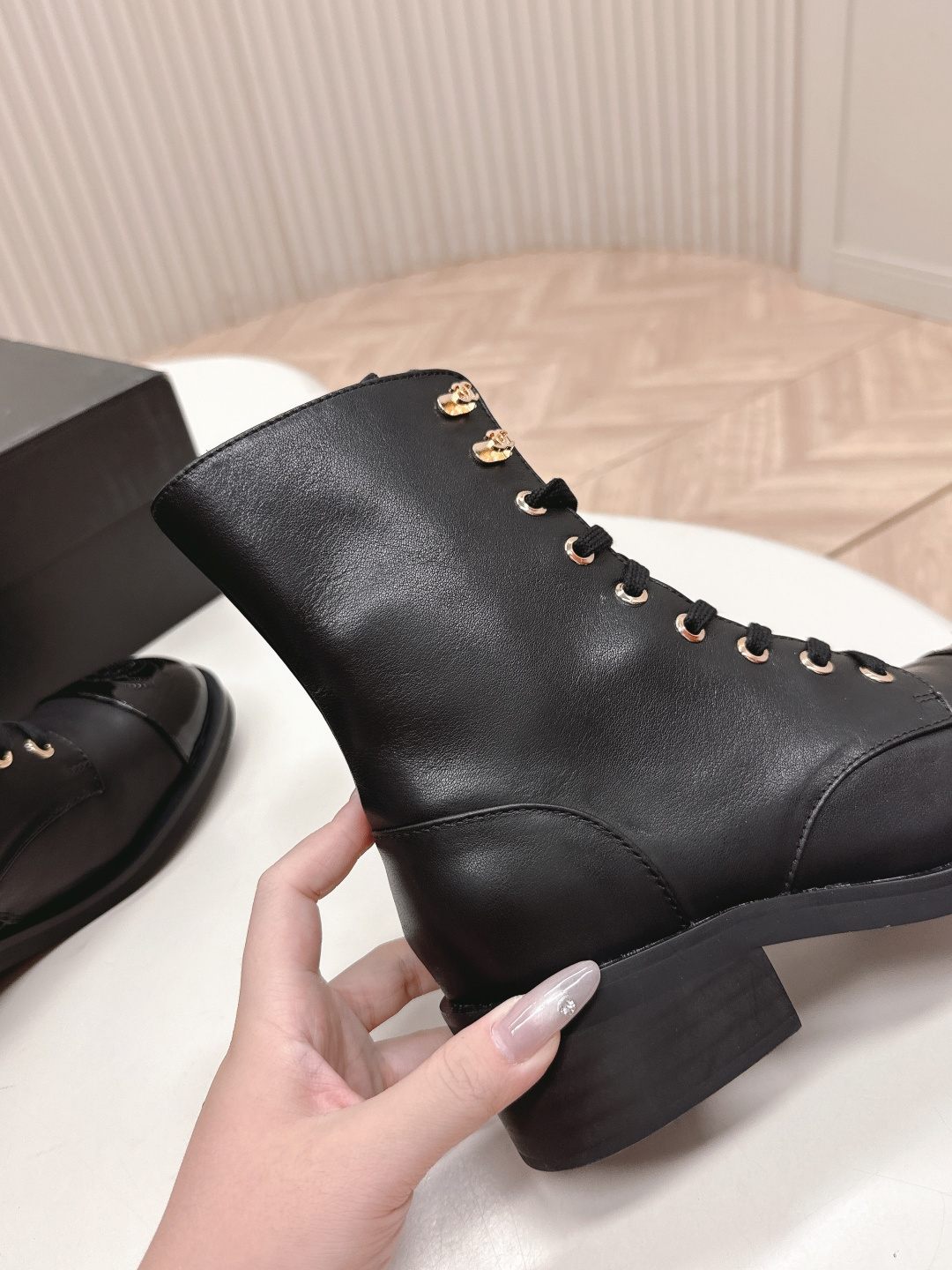 Pair of black leather lace-up combat boots with patent leather toe caps and gold hardware on a white table next to a black shoe box. - Hình 8