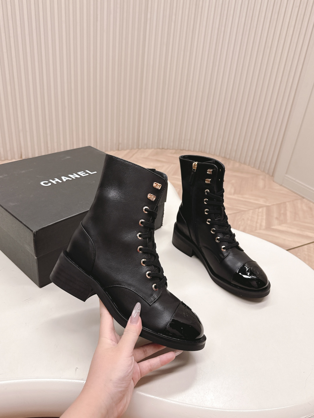 Pair of black leather lace-up combat boots with patent leather toe caps and gold hardware on a white table next to a black shoe box. - Hình 7