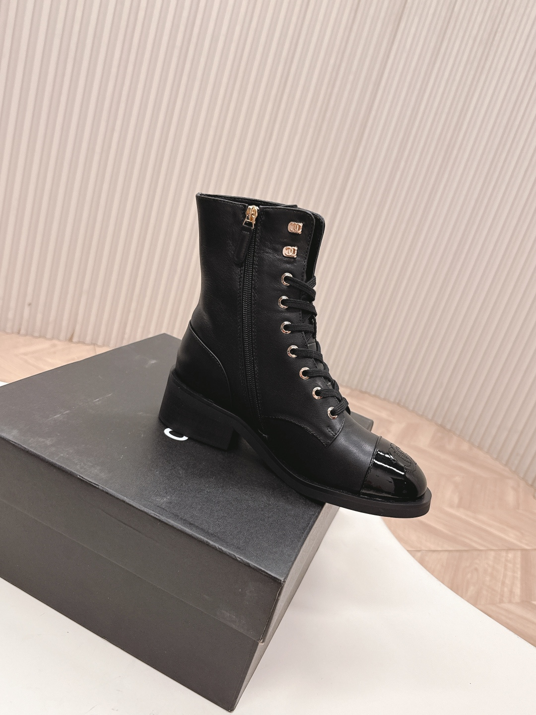 Pair of black leather lace-up combat boots with patent leather toe caps and gold hardware on a white table next to a black shoe box. - Hình 5