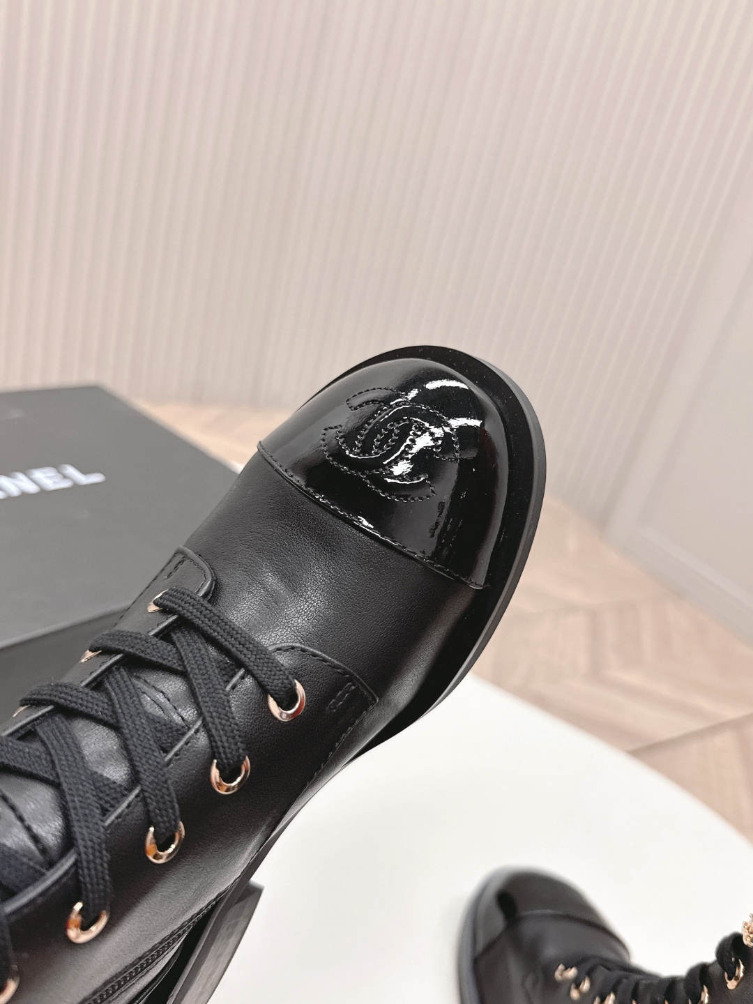 Pair of black leather lace-up combat boots with patent leather toe caps and gold hardware on a white table next to a black shoe box. - Hình 4