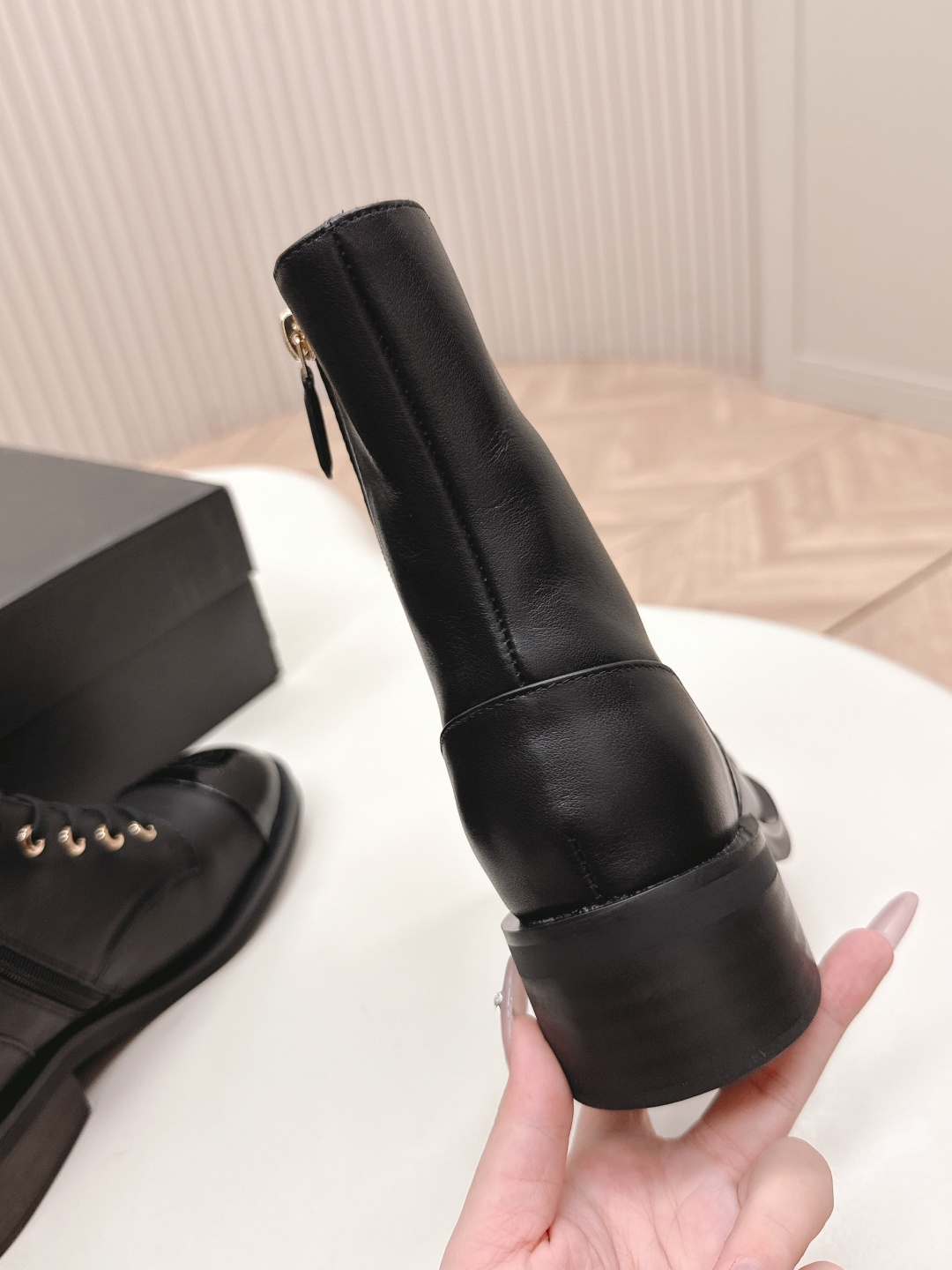 Pair of black leather lace-up combat boots with patent leather toe caps and gold hardware on a white table next to a black shoe box. - Hình 3