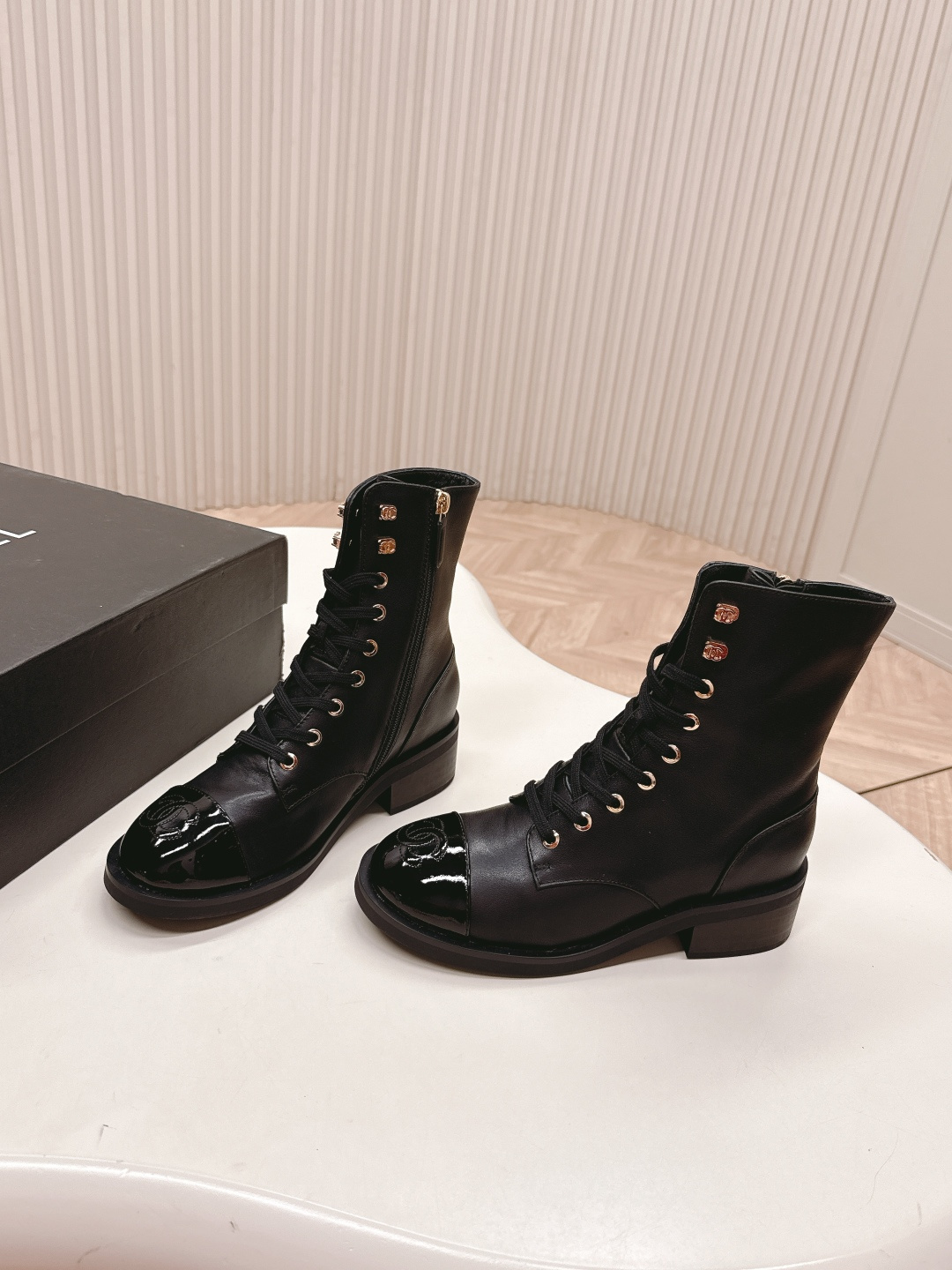 Pair of black leather lace-up combat boots with patent leather toe caps and gold hardware on a white table next to a black shoe box. - Hình 2