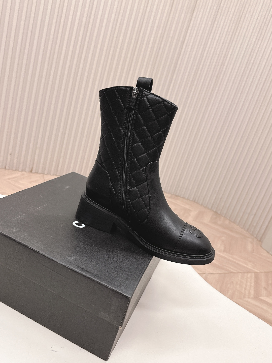 A pair of Chanel black quilted leather ankle boots with gold CC logos and side zippers placed on a white surface next to a black shoe box. - Hình 9