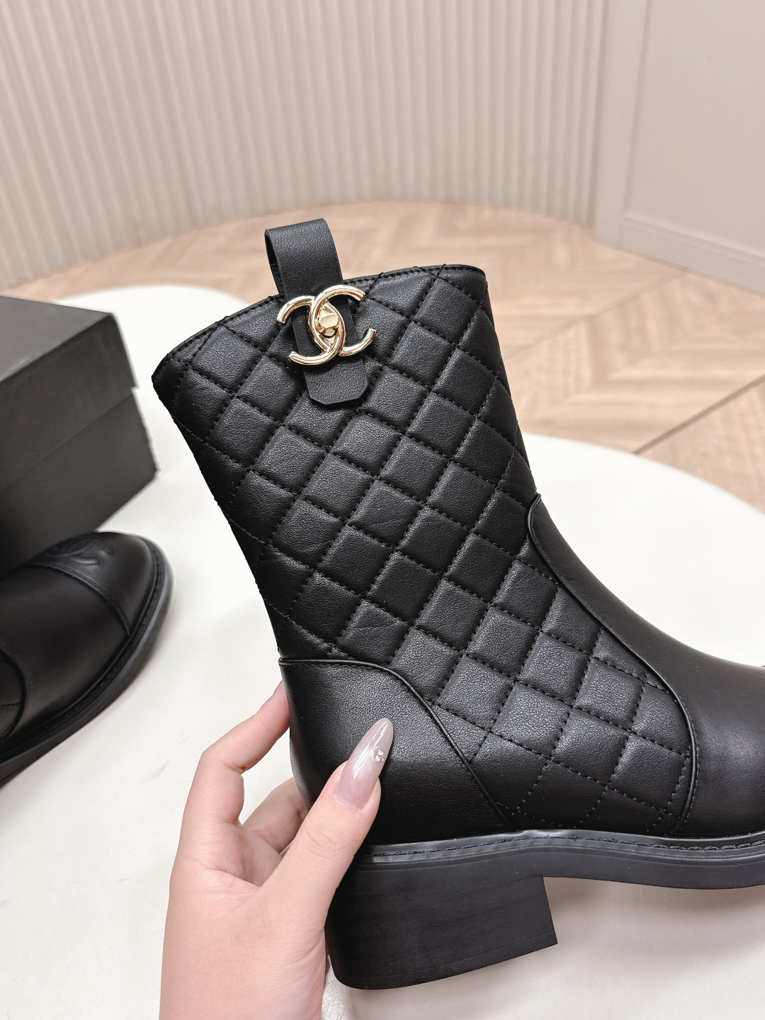 A pair of Chanel black quilted leather ankle boots with gold CC logos and side zippers placed on a white surface next to a black shoe box. - Hình 7