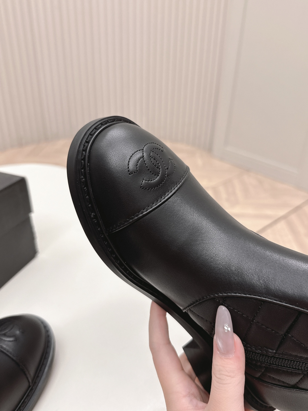 A pair of Chanel black quilted leather ankle boots with gold CC logos and side zippers placed on a white surface next to a black shoe box. - Hình 6
