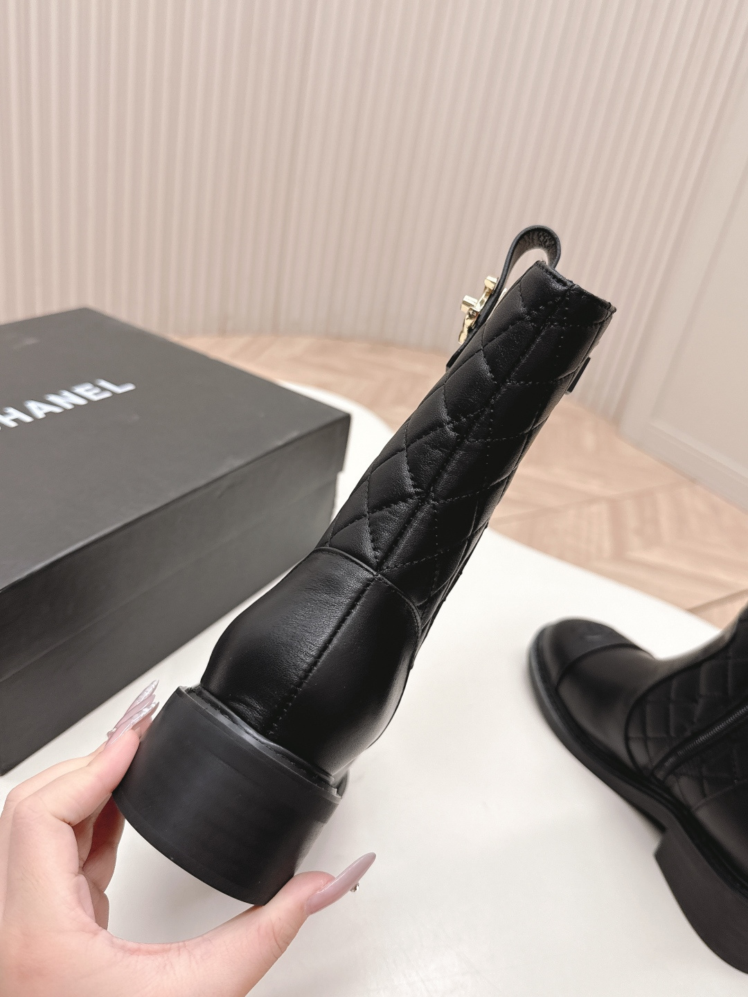 A pair of Chanel black quilted leather ankle boots with gold CC logos and side zippers placed on a white surface next to a black shoe box. - Hình 5