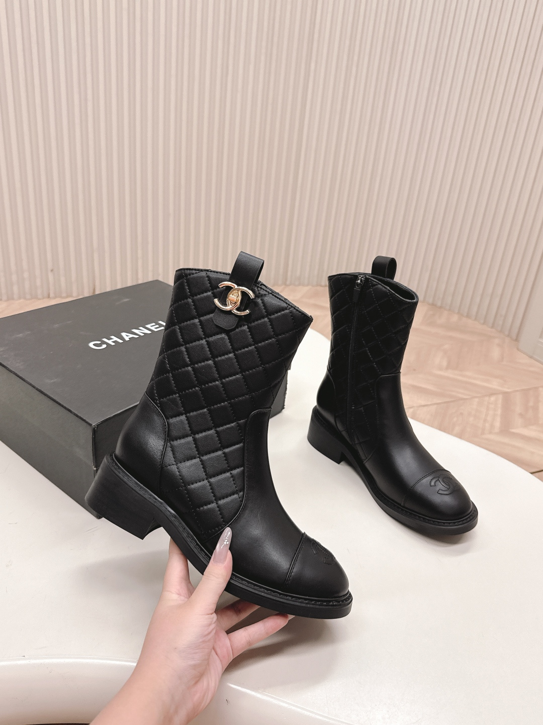 A pair of Chanel black quilted leather ankle boots with gold CC logos and side zippers placed on a white surface next to a black shoe box. - Hình 3