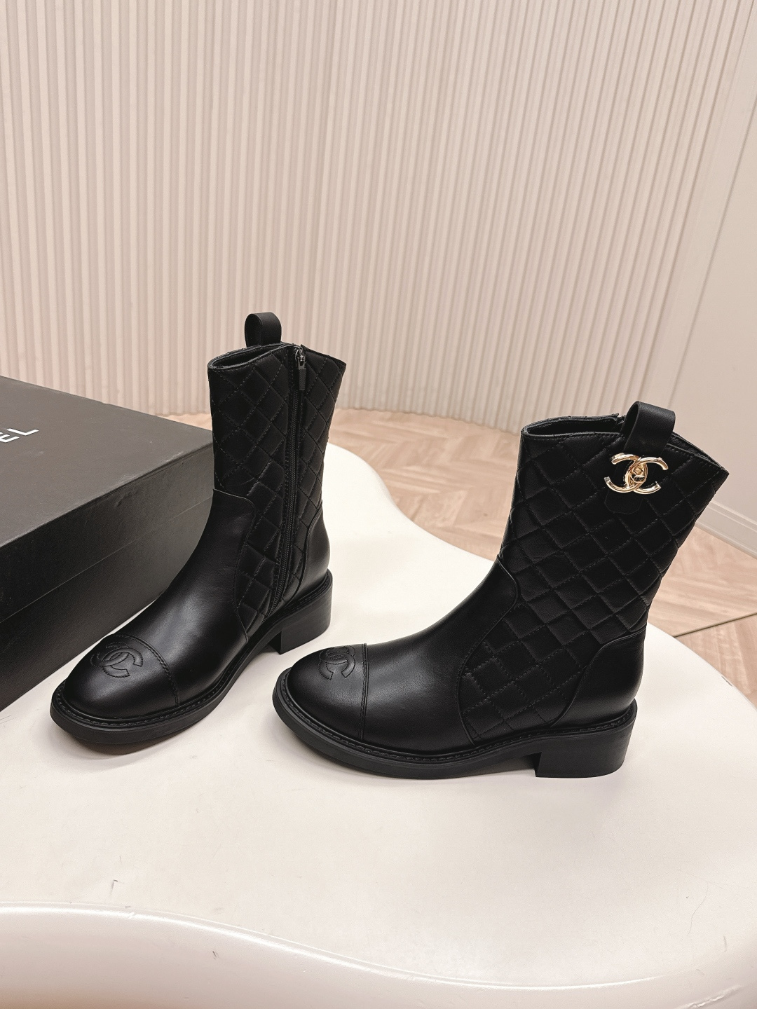 A pair of Chanel black quilted leather ankle boots with gold CC logos and side zippers placed on a white surface next to a black shoe box. - Hình 2