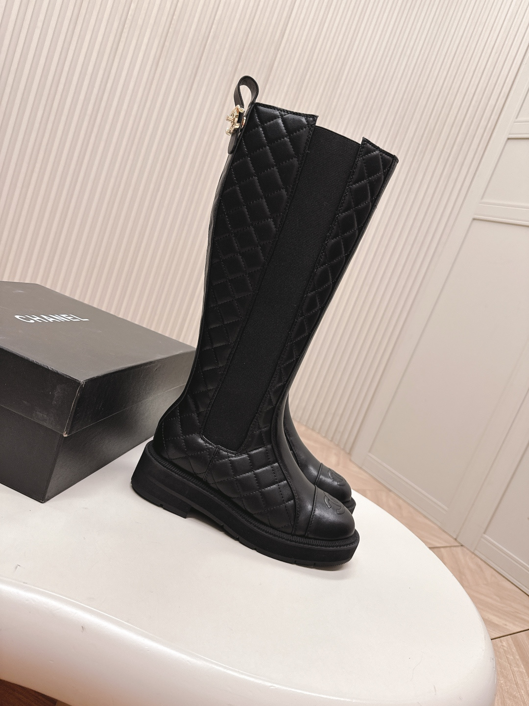 A pair of luxury Chanel black quilted leather knee-high boots with the CC logo on the toe cap and a chunky lug sole, displayed next to a Chanel shoe box. - Hình 8