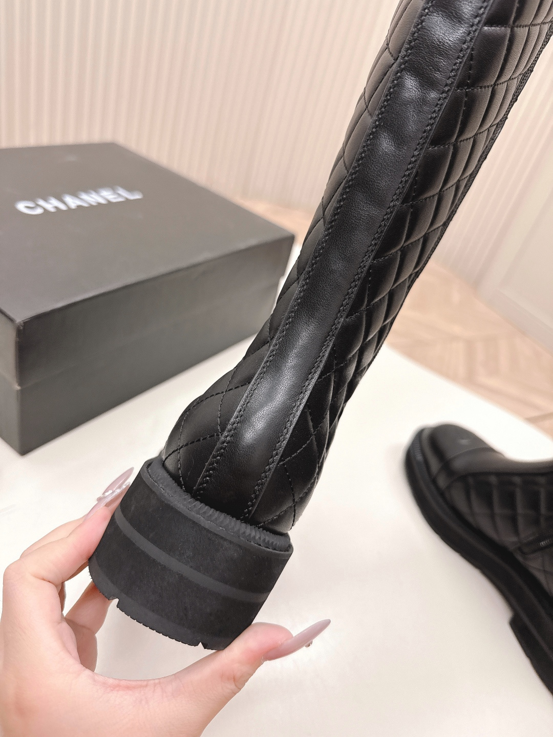 A pair of luxury Chanel black quilted leather knee-high boots with the CC logo on the toe cap and a chunky lug sole, displayed next to a Chanel shoe box. - Hình 7