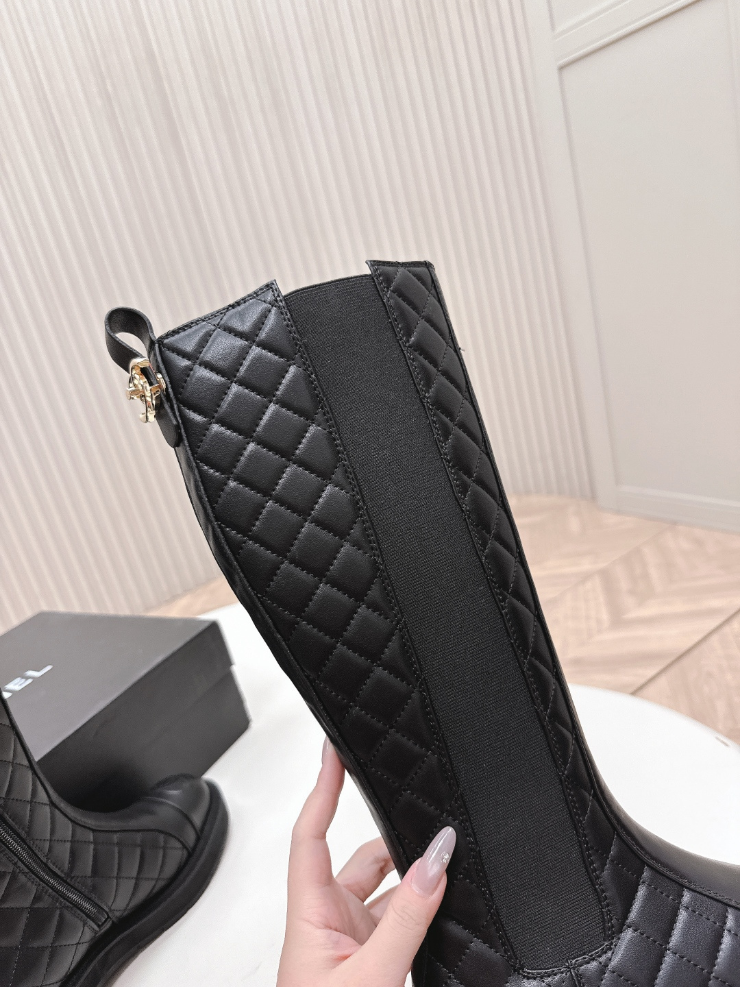 A pair of luxury Chanel black quilted leather knee-high boots with the CC logo on the toe cap and a chunky lug sole, displayed next to a Chanel shoe box. - Hình 6