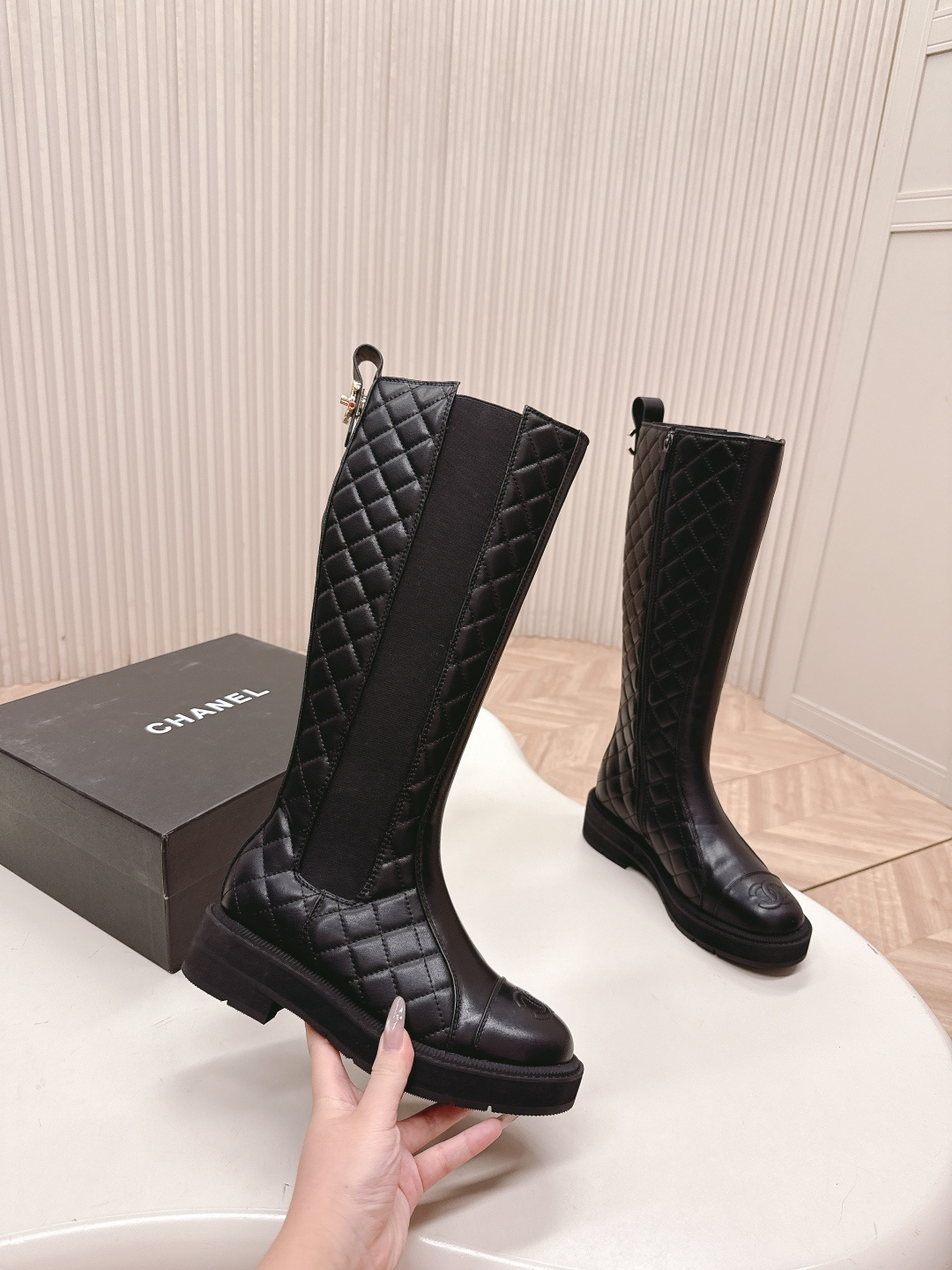 A pair of luxury Chanel black quilted leather knee-high boots with the CC logo on the toe cap and a chunky lug sole, displayed next to a Chanel shoe box. - Hình 4