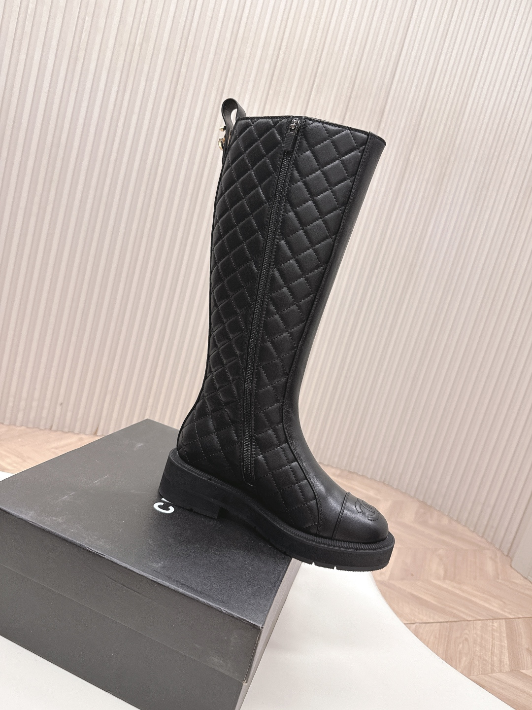 A pair of luxury Chanel black quilted leather knee-high boots with the CC logo on the toe cap and a chunky lug sole, displayed next to a Chanel shoe box. - Hình 3