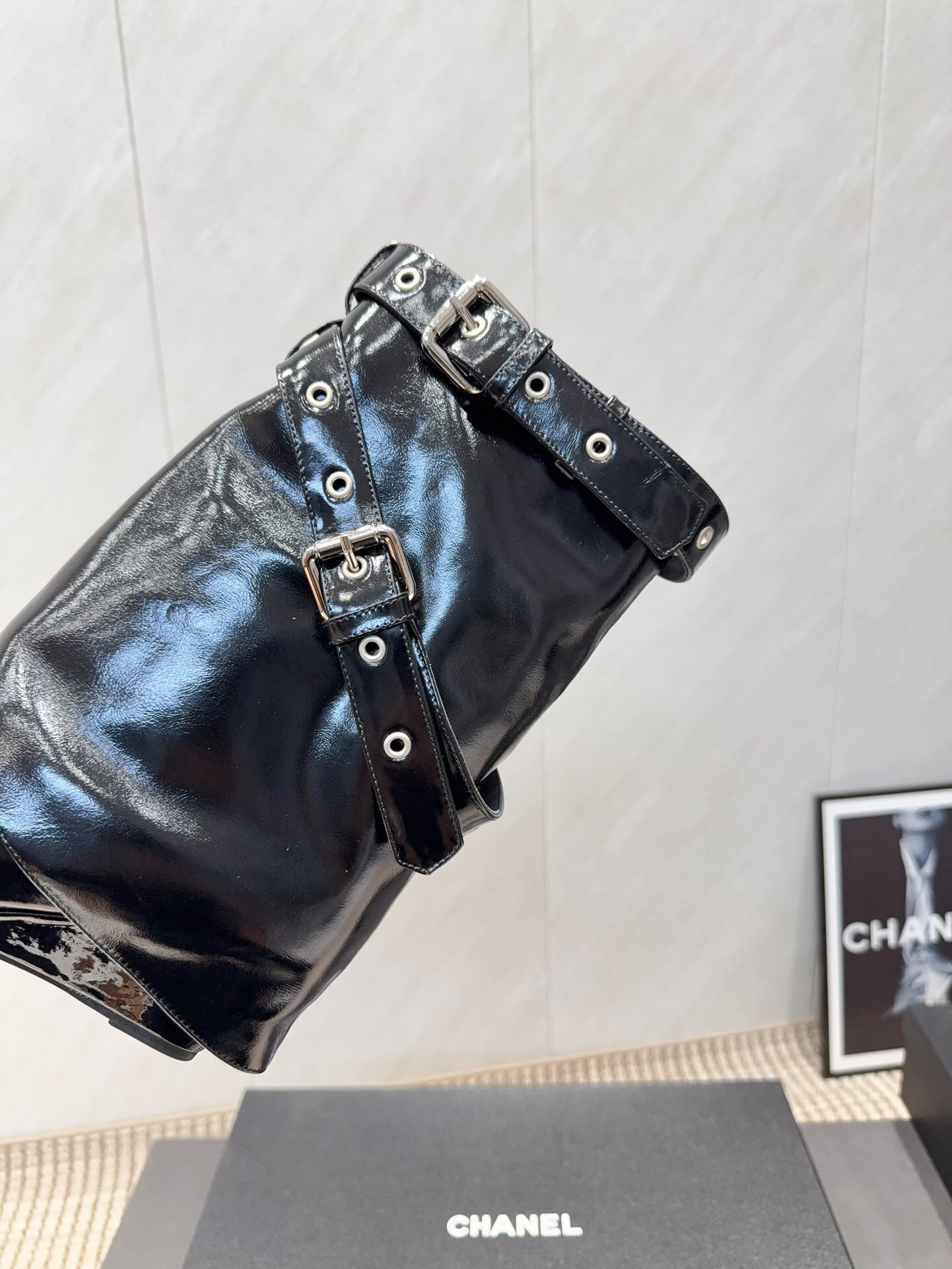A pair of shiny black Chanel patent leather ankle boots featuring multiple silver buckle straps and a sharp pointed toe sitting on a designer box. - Hình 7