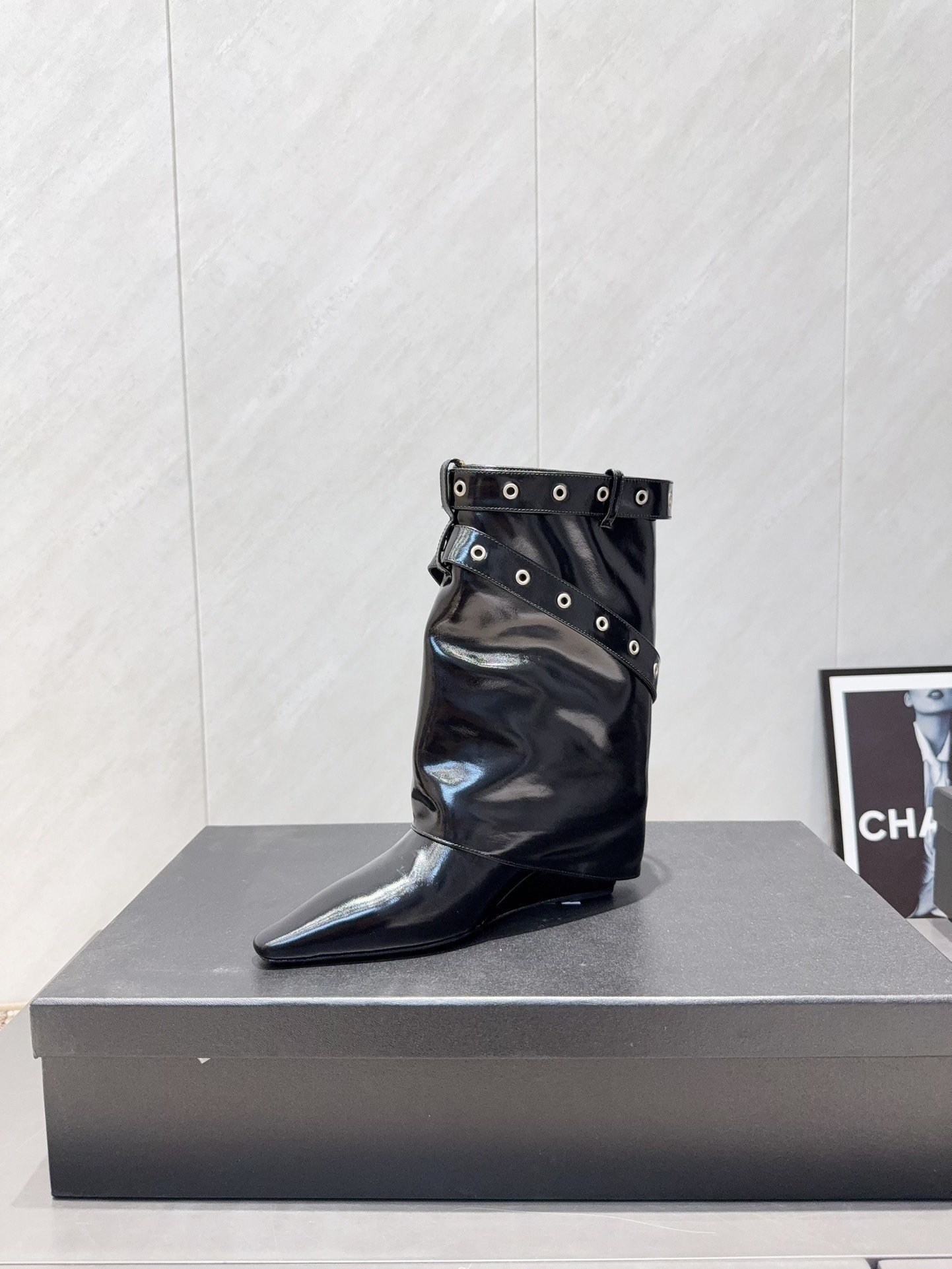 A pair of shiny black Chanel patent leather ankle boots featuring multiple silver buckle straps and a sharp pointed toe sitting on a designer box. - Hình 6