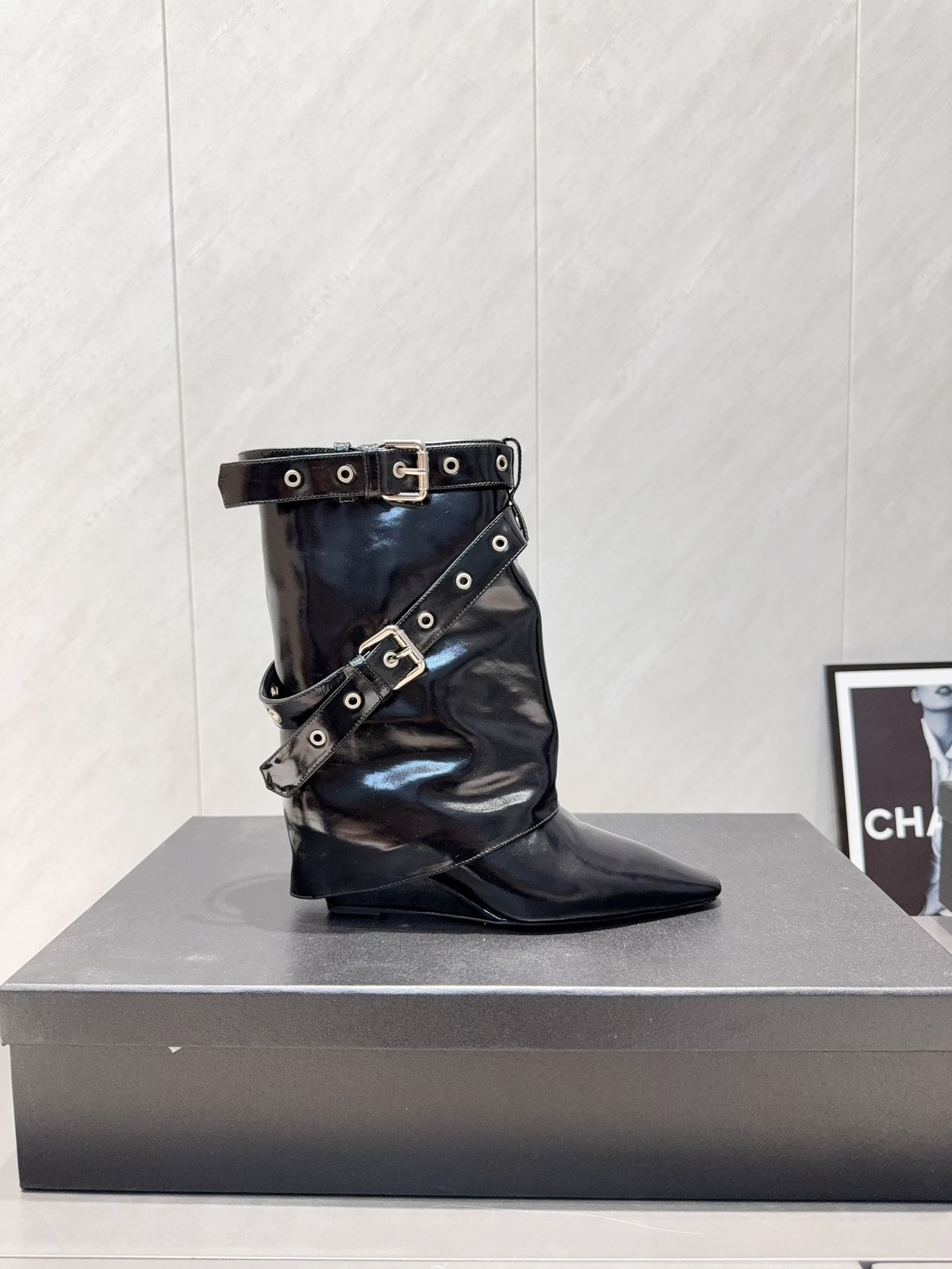 A pair of shiny black Chanel patent leather ankle boots featuring multiple silver buckle straps and a sharp pointed toe sitting on a designer box. - Hình 5