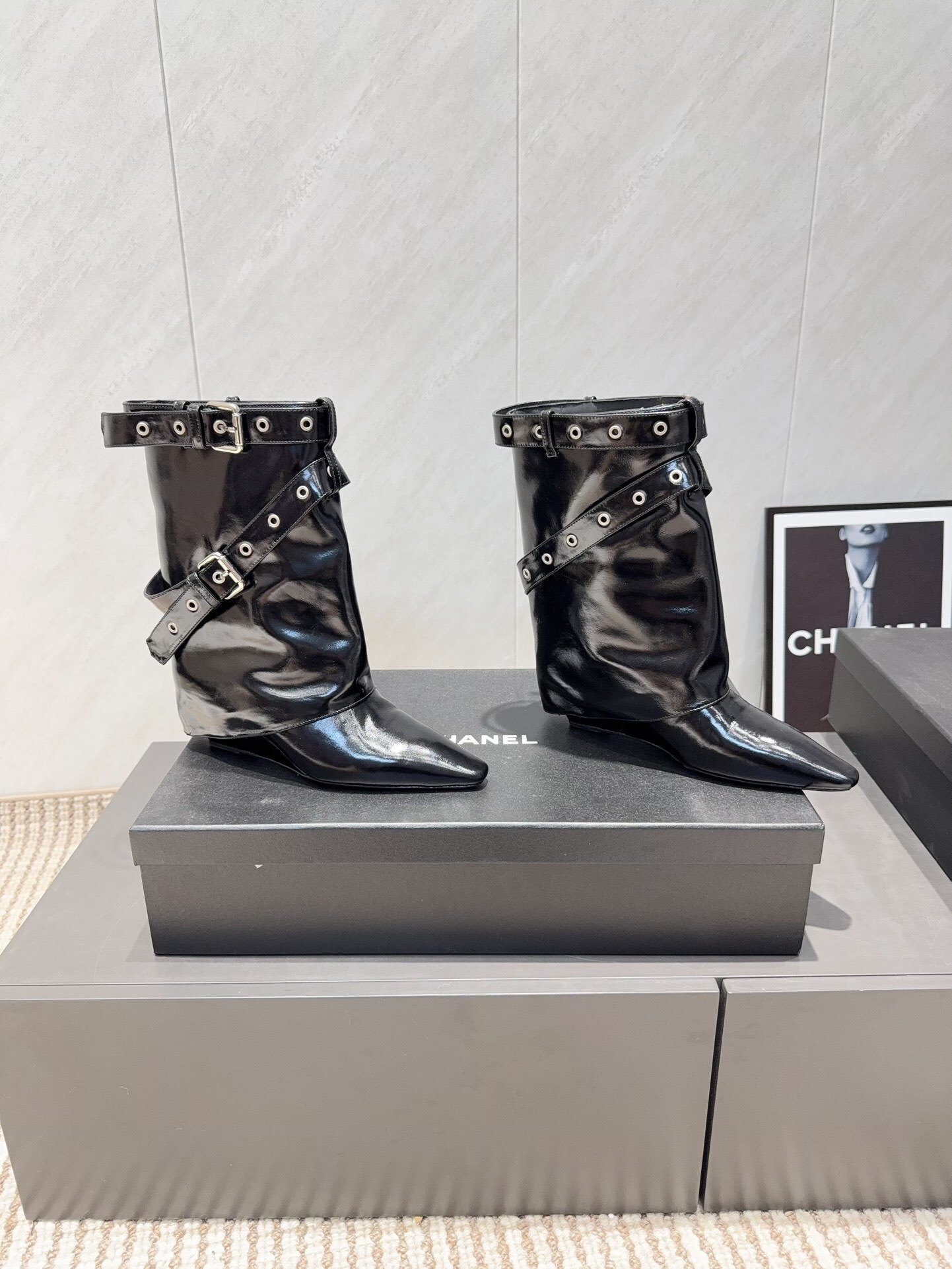 A pair of shiny black Chanel patent leather ankle boots featuring multiple silver buckle straps and a sharp pointed toe sitting on a designer box. - Hình 3