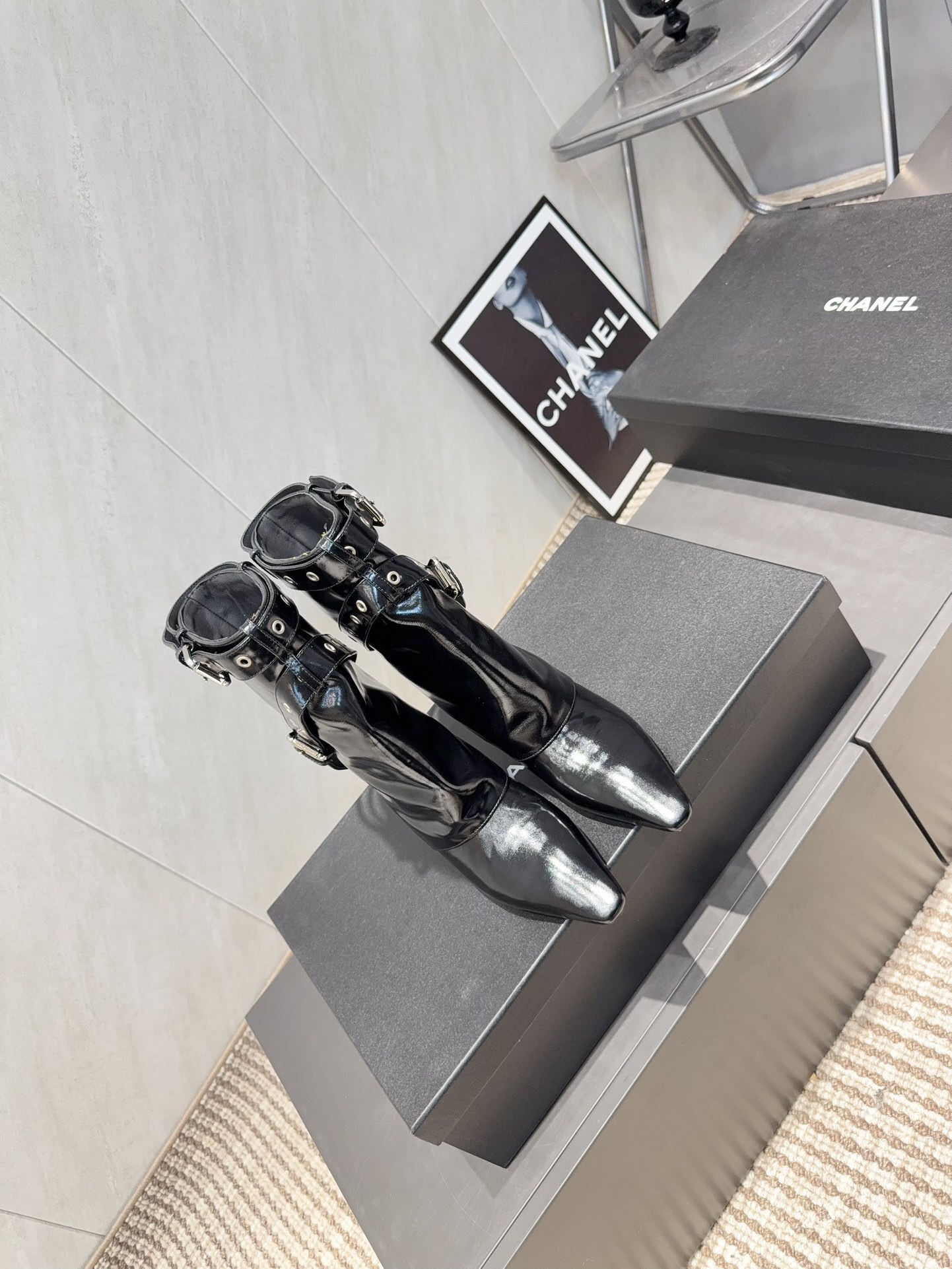 A pair of shiny black Chanel patent leather ankle boots featuring multiple silver buckle straps and a sharp pointed toe sitting on a designer box. - Hình 2