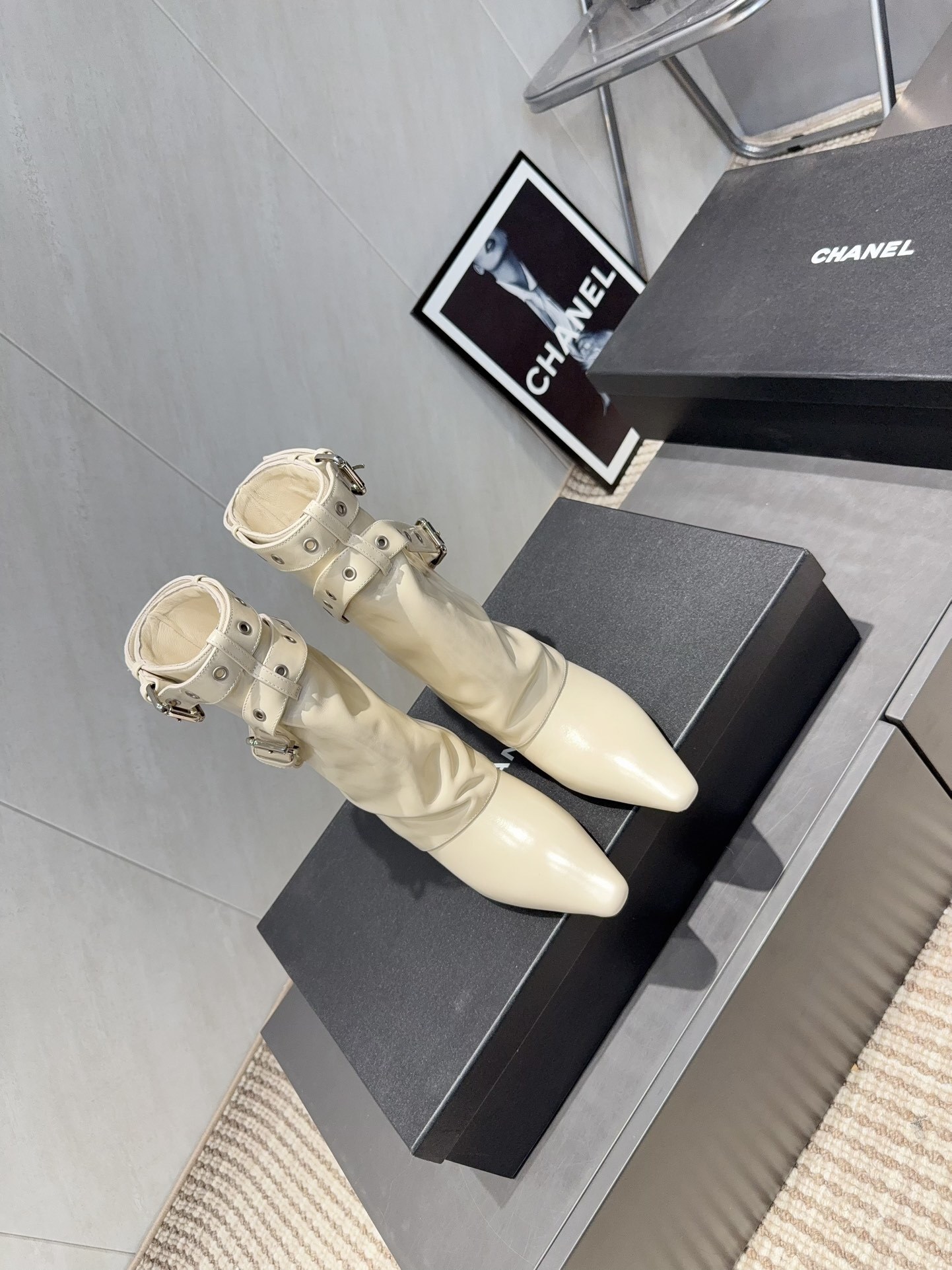 A pair of Chanel cream leather slouchy ankle boots with silver buckle straps and pointed toes placed on a black branded box. - Hình 2