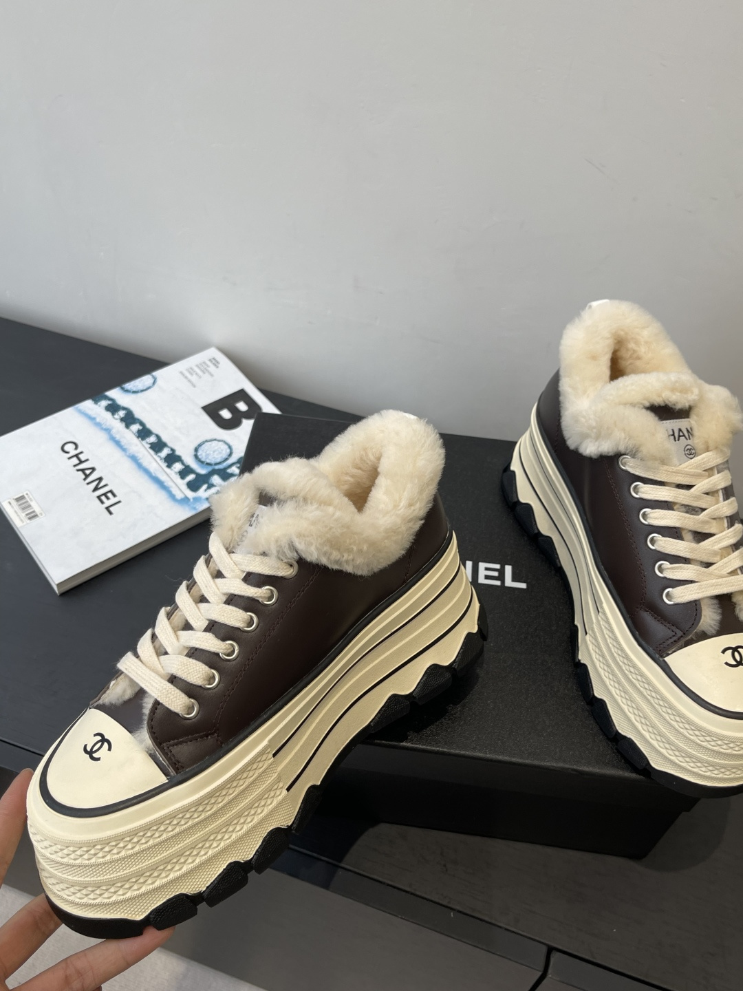 A pair of Chanel brown leather platform sneakers with cream soles and white shearling lining displayed on a black box. - Hình 3