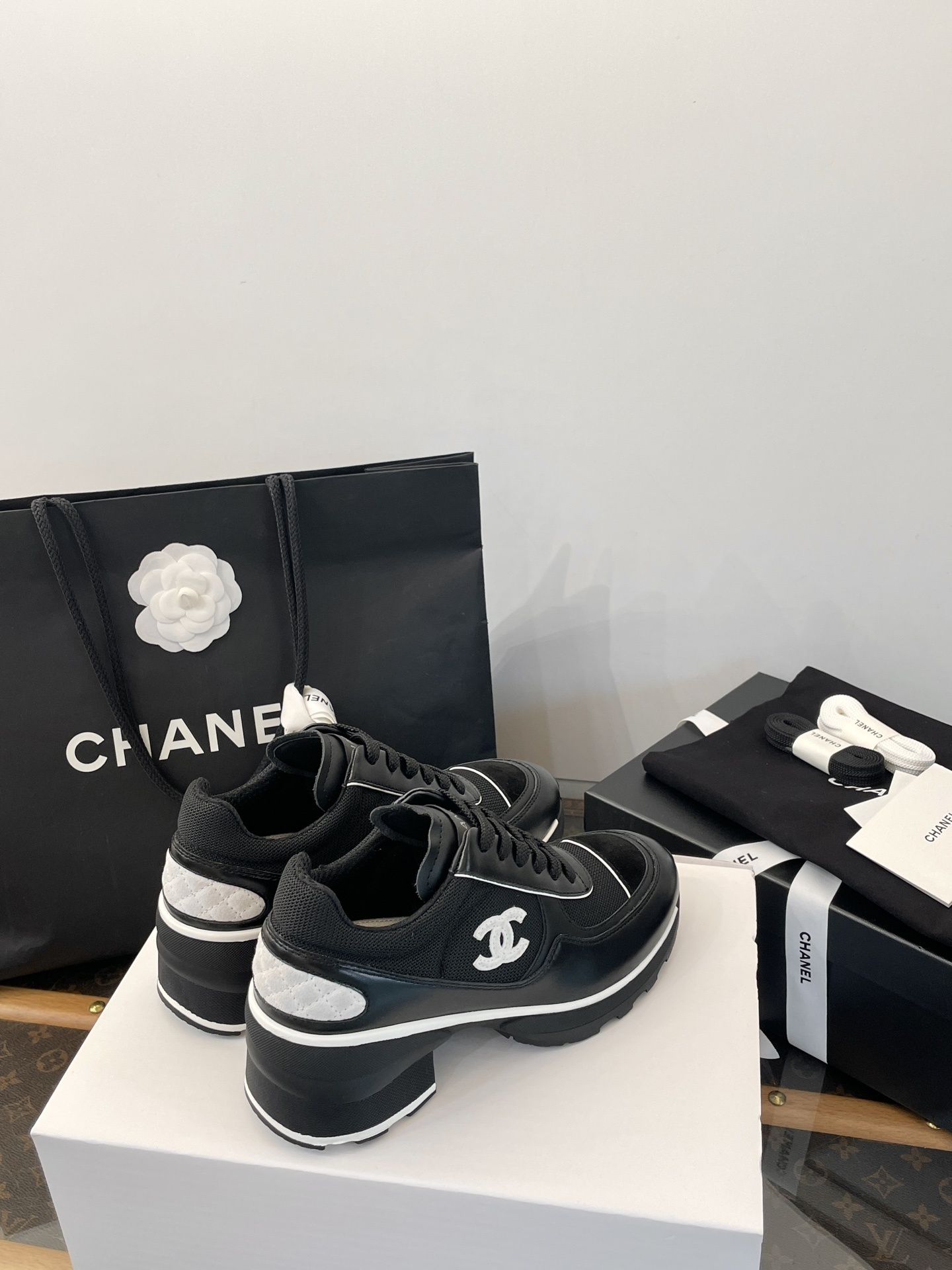 A pair of black Chanel sneakers with white CC logo on a white box with luxury Chanel packaging in the background. - Hình 7