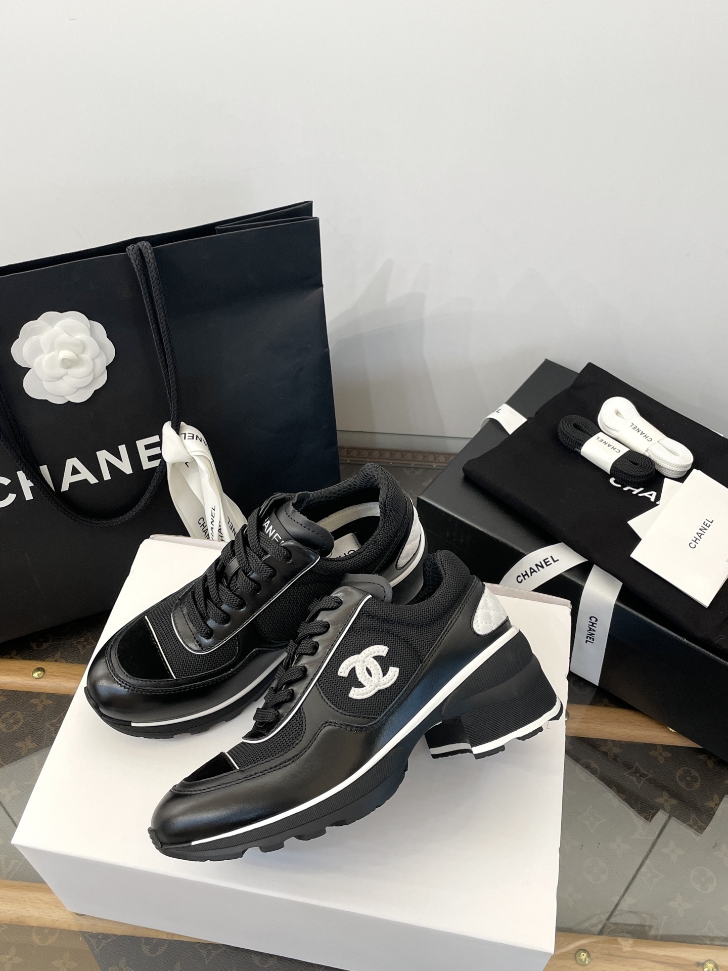 A pair of black Chanel sneakers with white CC logo on a white box with luxury Chanel packaging in the background. - Hình 6