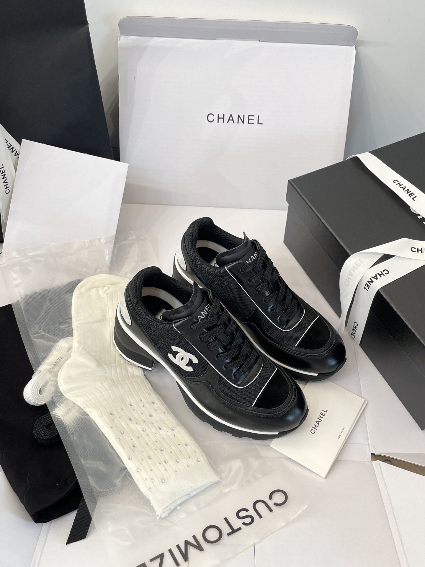 A pair of black Chanel sneakers with white CC logo on a white box with luxury Chanel packaging in the background. - Hình 5