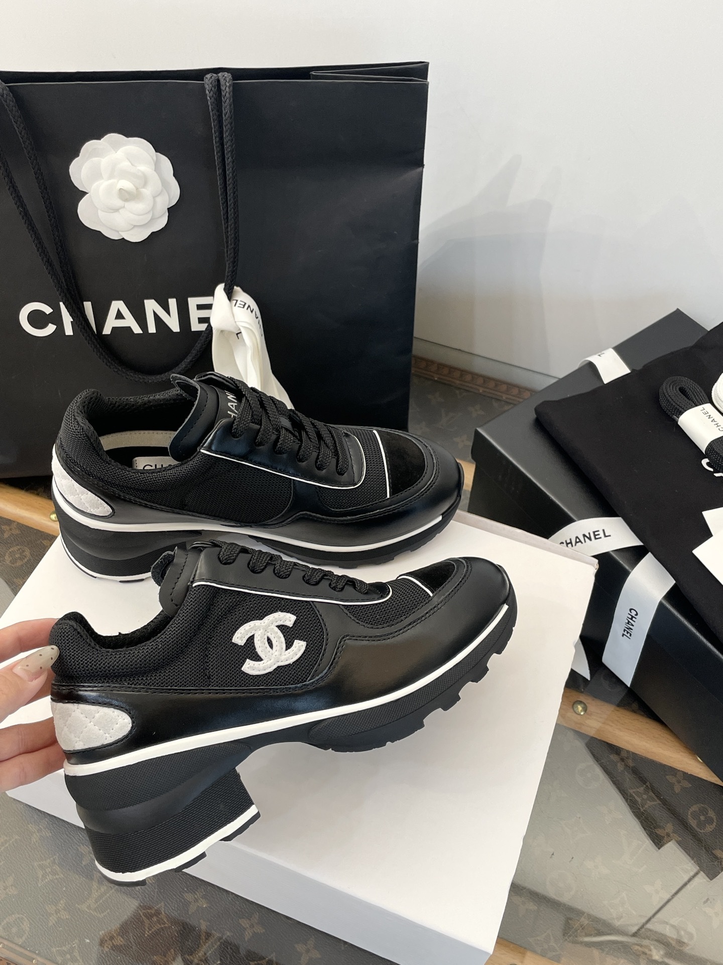 A pair of black Chanel sneakers with white CC logo on a white box with luxury Chanel packaging in the background. - Hình 4