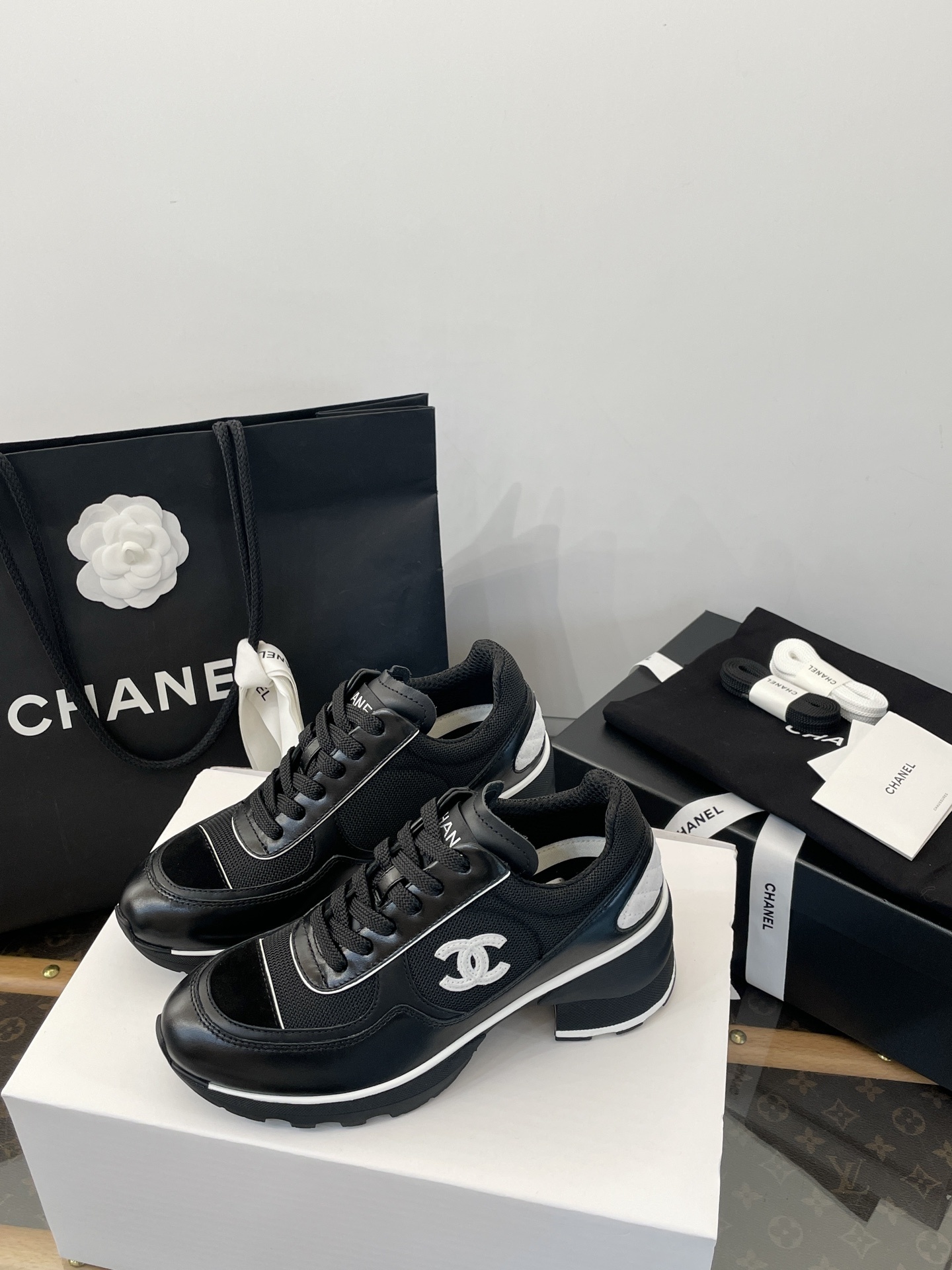 A pair of black Chanel sneakers with white CC logo on a white box with luxury Chanel packaging in the background. - Hình 3