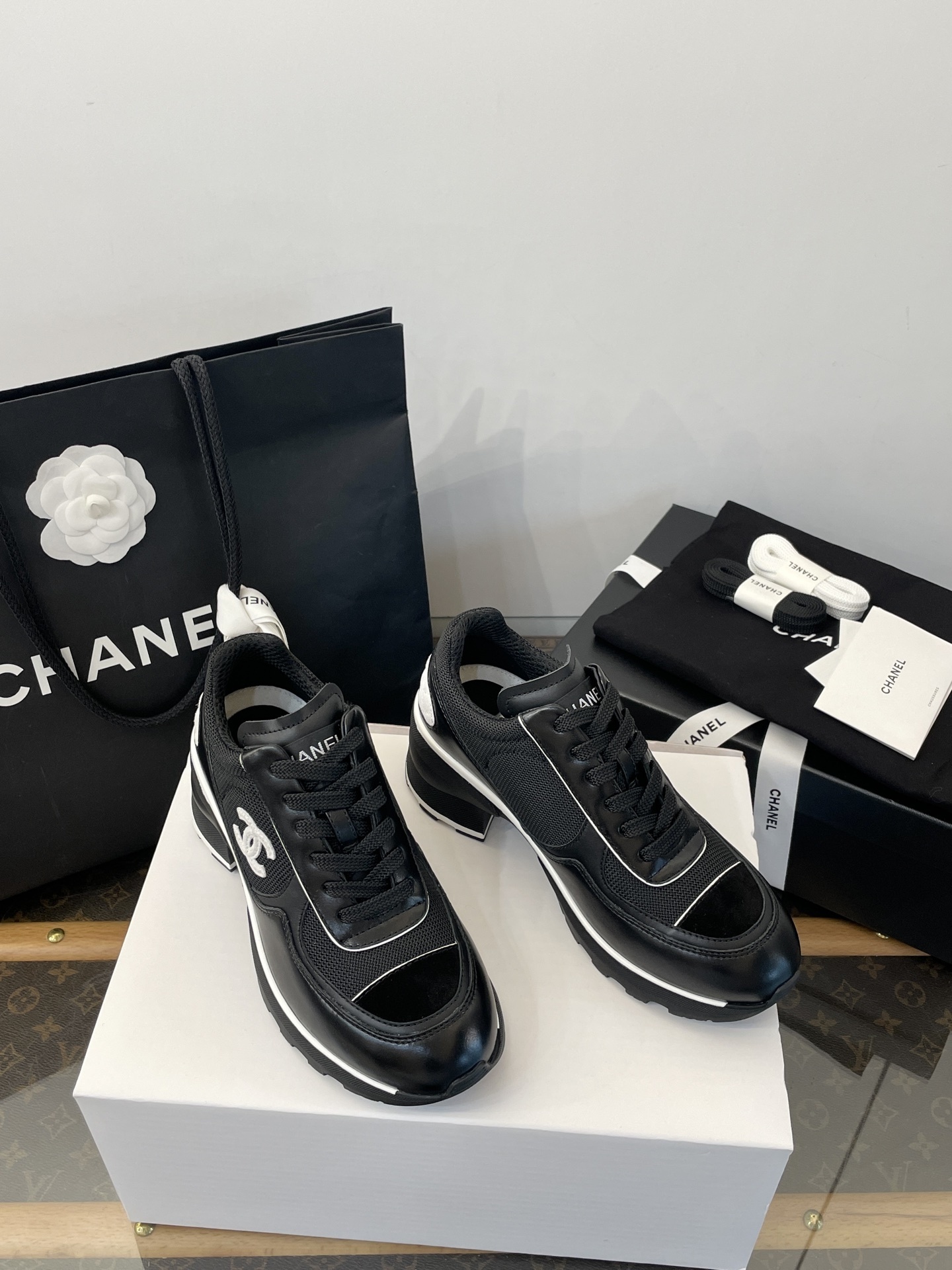A pair of black Chanel sneakers with white CC logo on a white box with luxury Chanel packaging in the background. - Hình 2