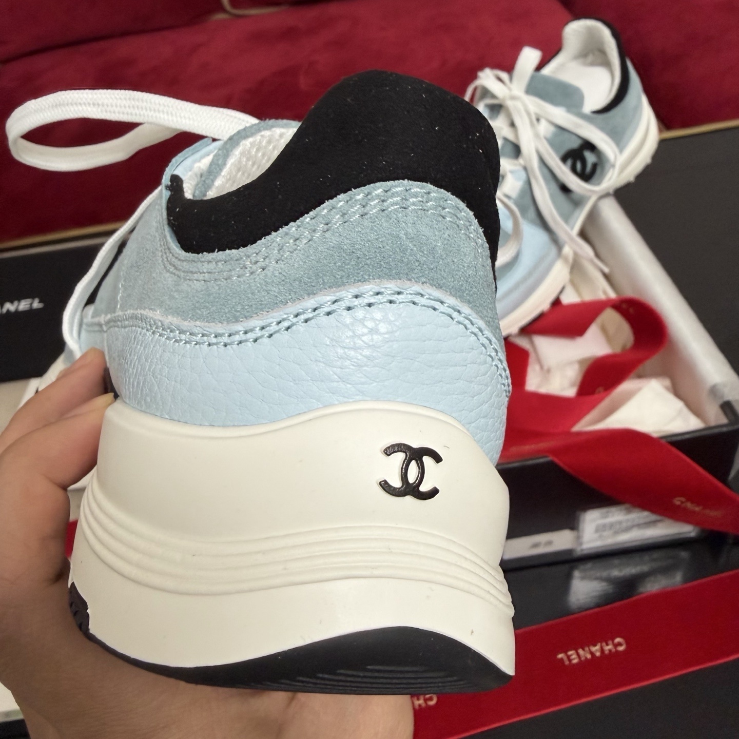A pair of light blue Chanel sneakers with a black CC logo and white laces displayed with luxury packaging and red ribbons. - Hình 8