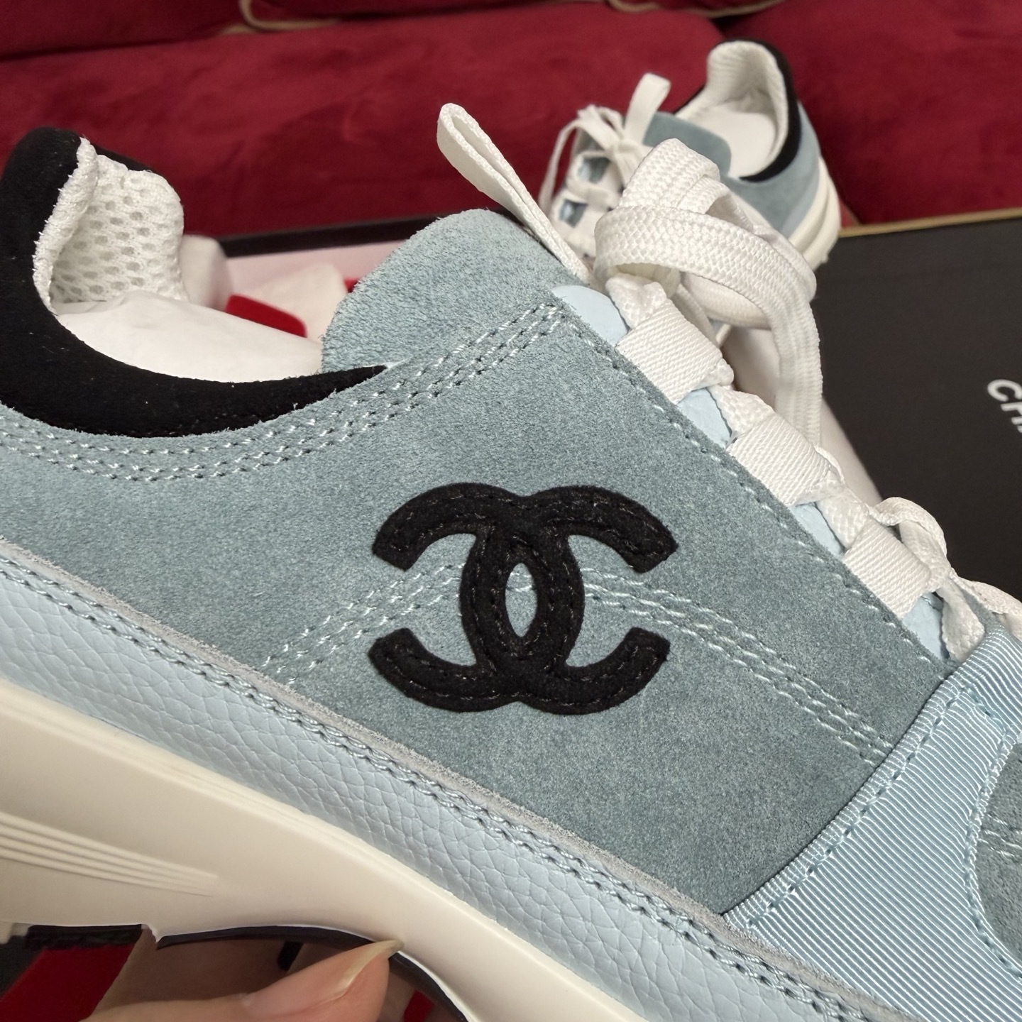 A pair of light blue Chanel sneakers with a black CC logo and white laces displayed with luxury packaging and red ribbons. - Hình 7