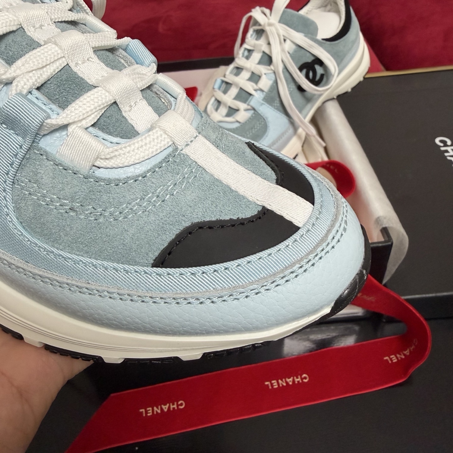 A pair of light blue Chanel sneakers with a black CC logo and white laces displayed with luxury packaging and red ribbons. - Hình 6