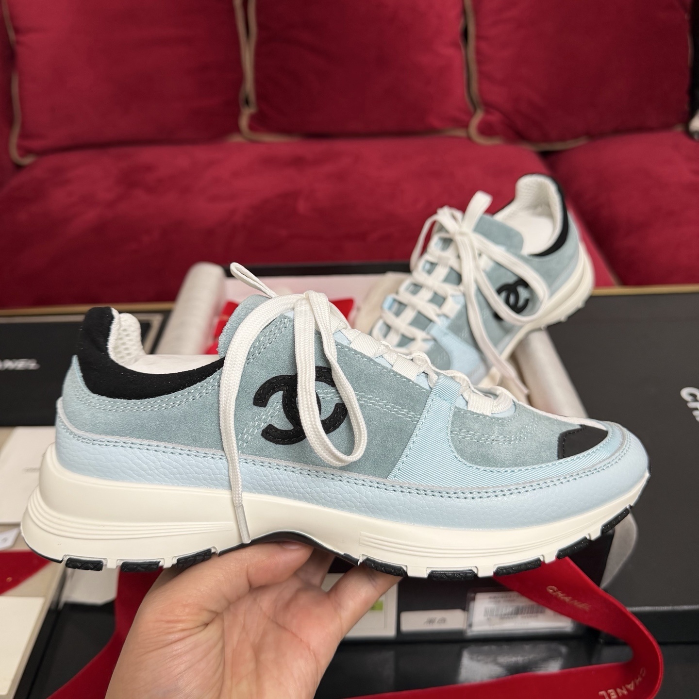 A pair of light blue Chanel sneakers with a black CC logo and white laces displayed with luxury packaging and red ribbons. - Hình 5