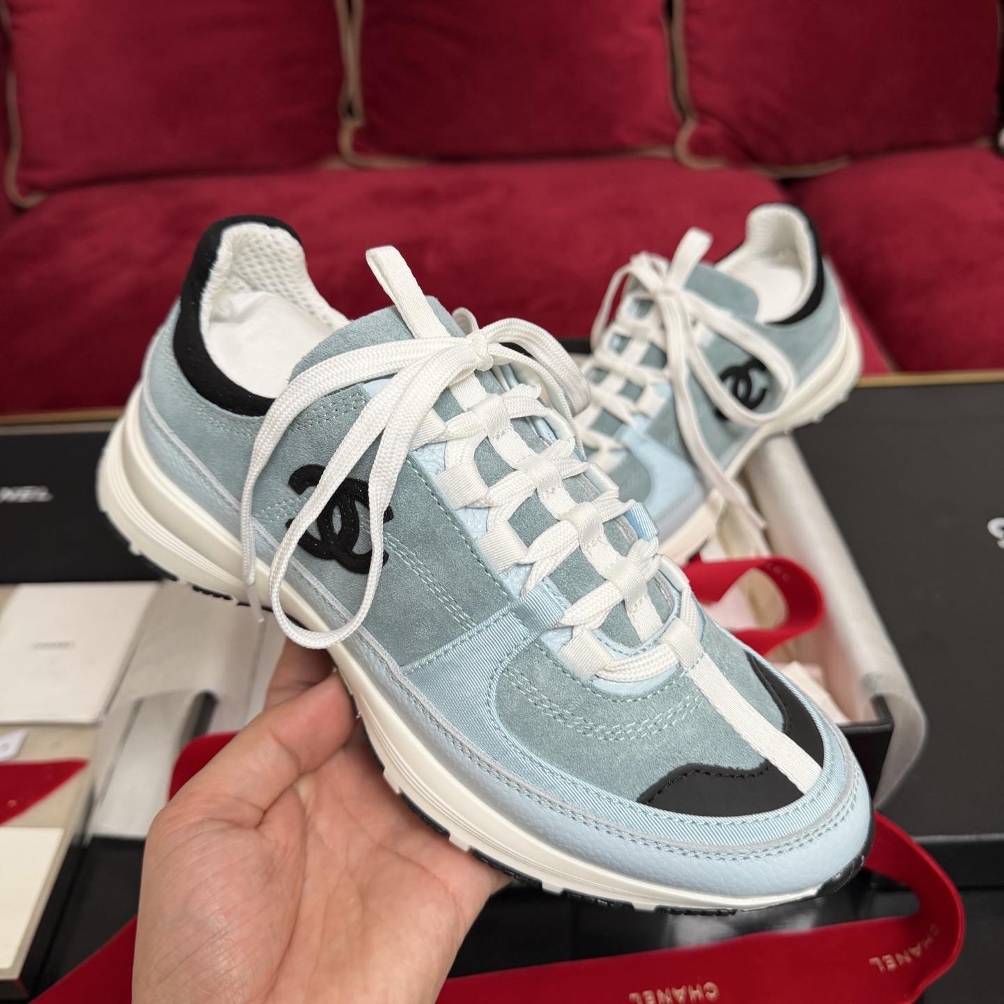A pair of light blue Chanel sneakers with a black CC logo and white laces displayed with luxury packaging and red ribbons. - Hình 4