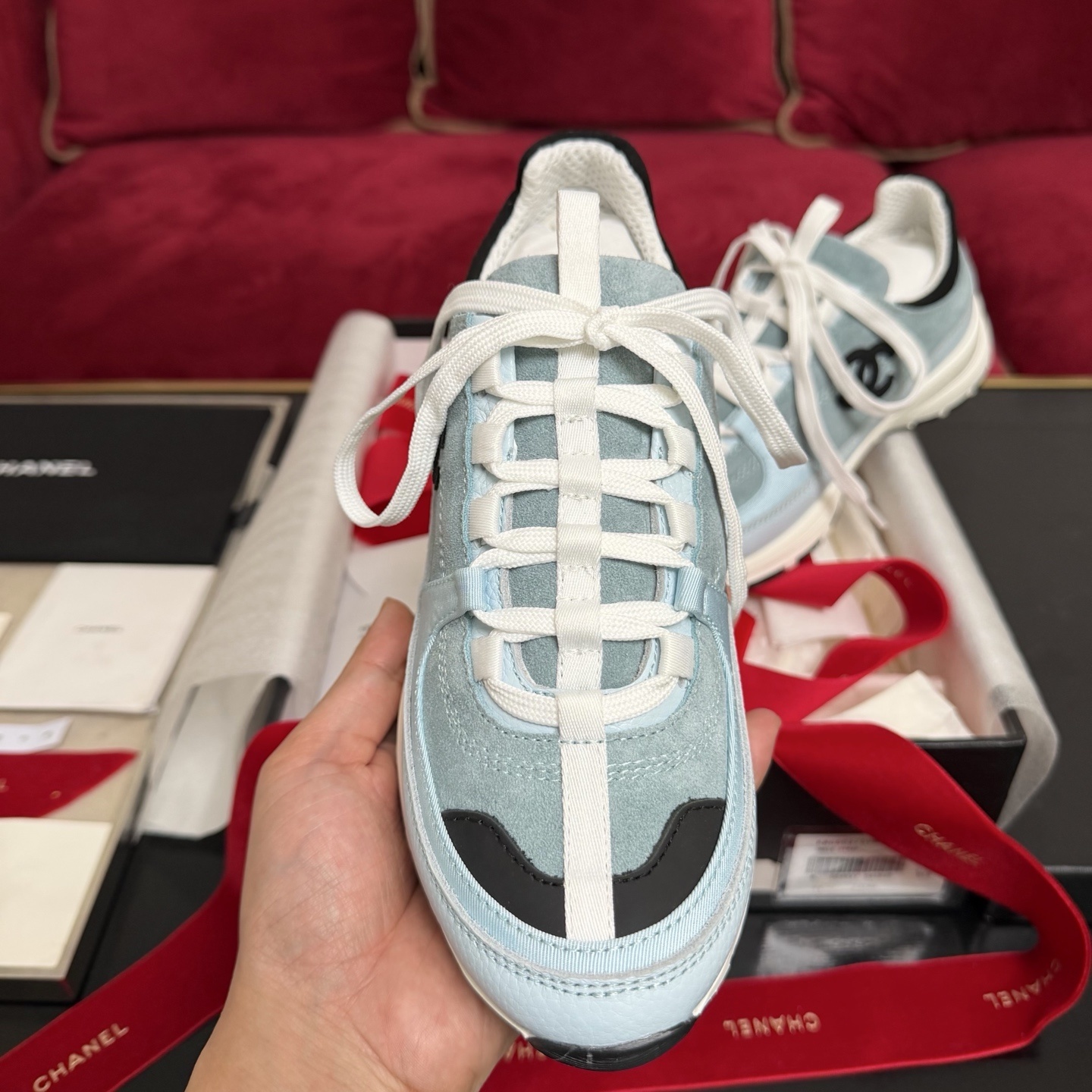 A pair of light blue Chanel sneakers with a black CC logo and white laces displayed with luxury packaging and red ribbons. - Hình 3
