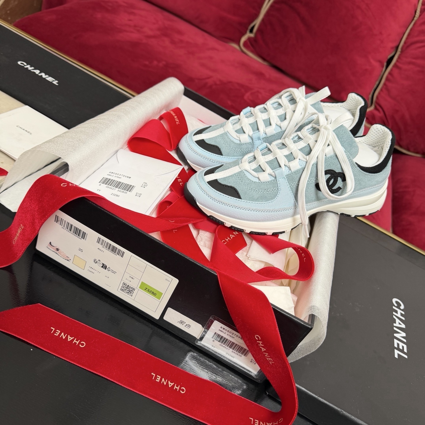 A pair of light blue Chanel sneakers with a black CC logo and white laces displayed with luxury packaging and red ribbons. - Hình 2
