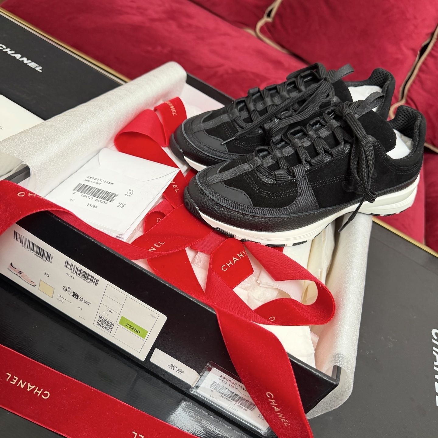 A pair of black Chanel suede and fabric sneakers sitting on a designer box with red ribbons. - Hình 2