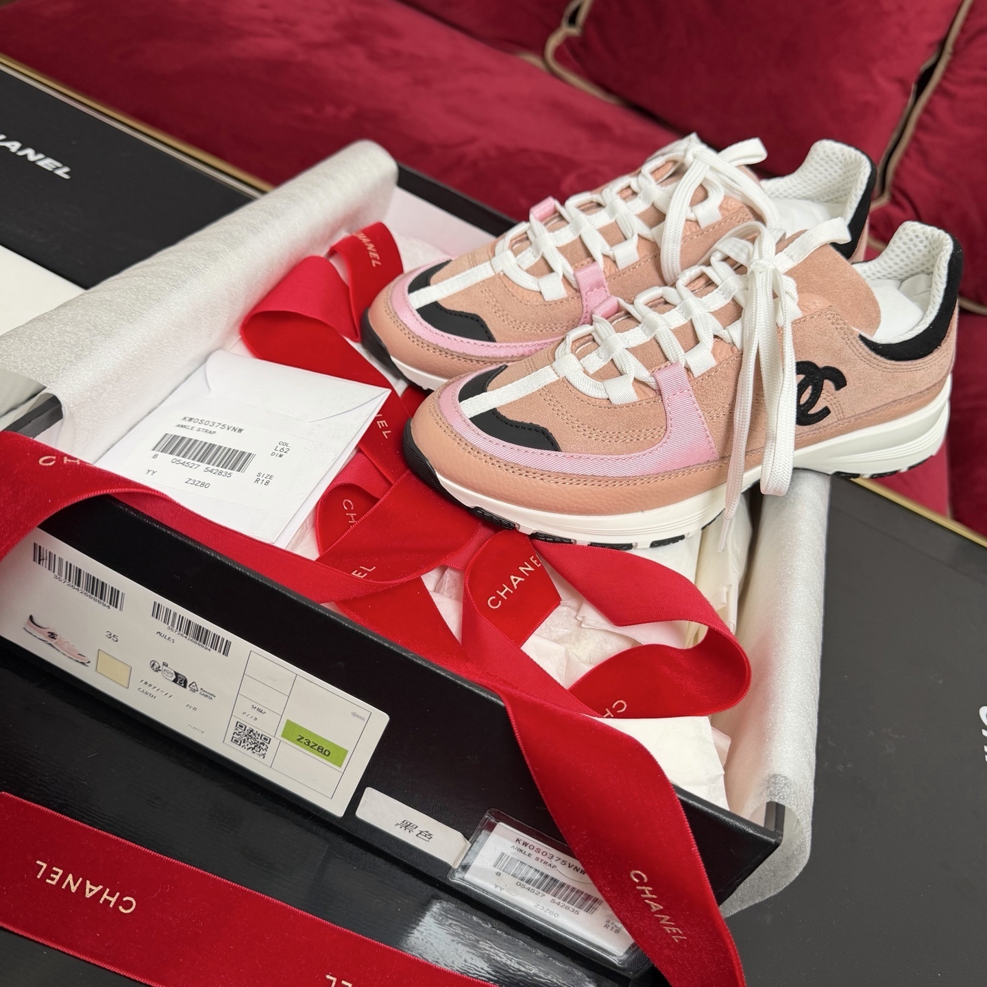 A pair of pink and black Chanel sneakers with white laces sitting on a luxury gift box with red ribbons - Hình 2