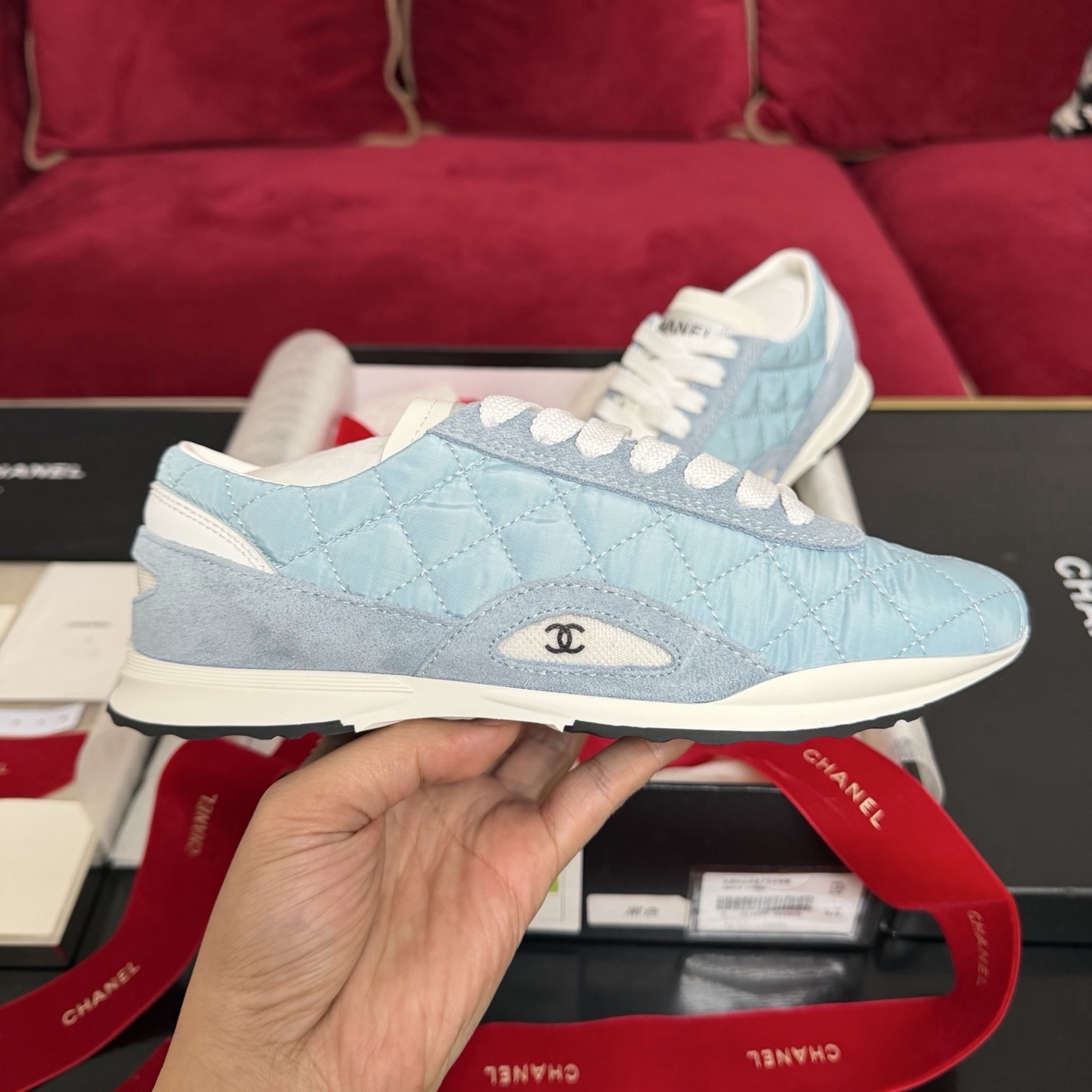 A pair of light blue quilted Chanel sneakers displayed with a black box, red ribbon, and authenticity cards. - Hình 5