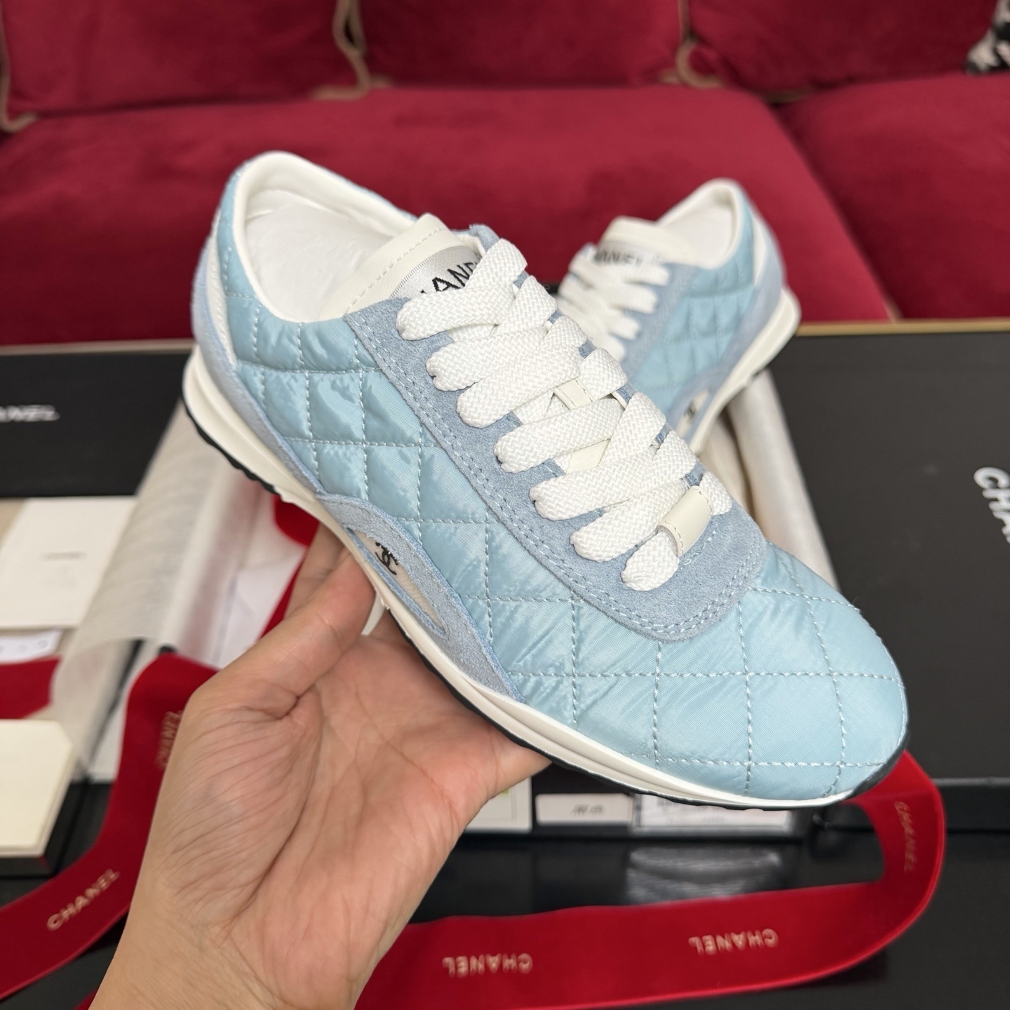 A pair of light blue quilted Chanel sneakers displayed with a black box, red ribbon, and authenticity cards. - Hình 4