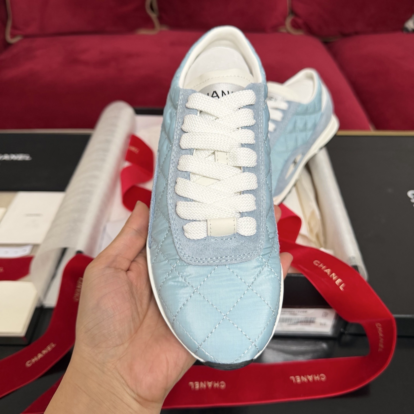 A pair of light blue quilted Chanel sneakers displayed with a black box, red ribbon, and authenticity cards. - Hình 3