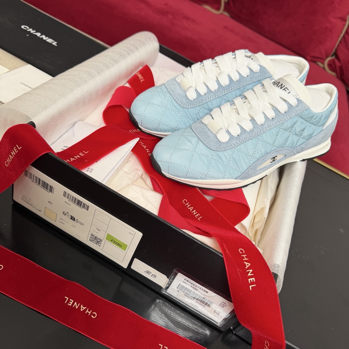 A pair of light blue quilted Chanel sneakers displayed with a black box, red ribbon, and authenticity cards. - Hình 2