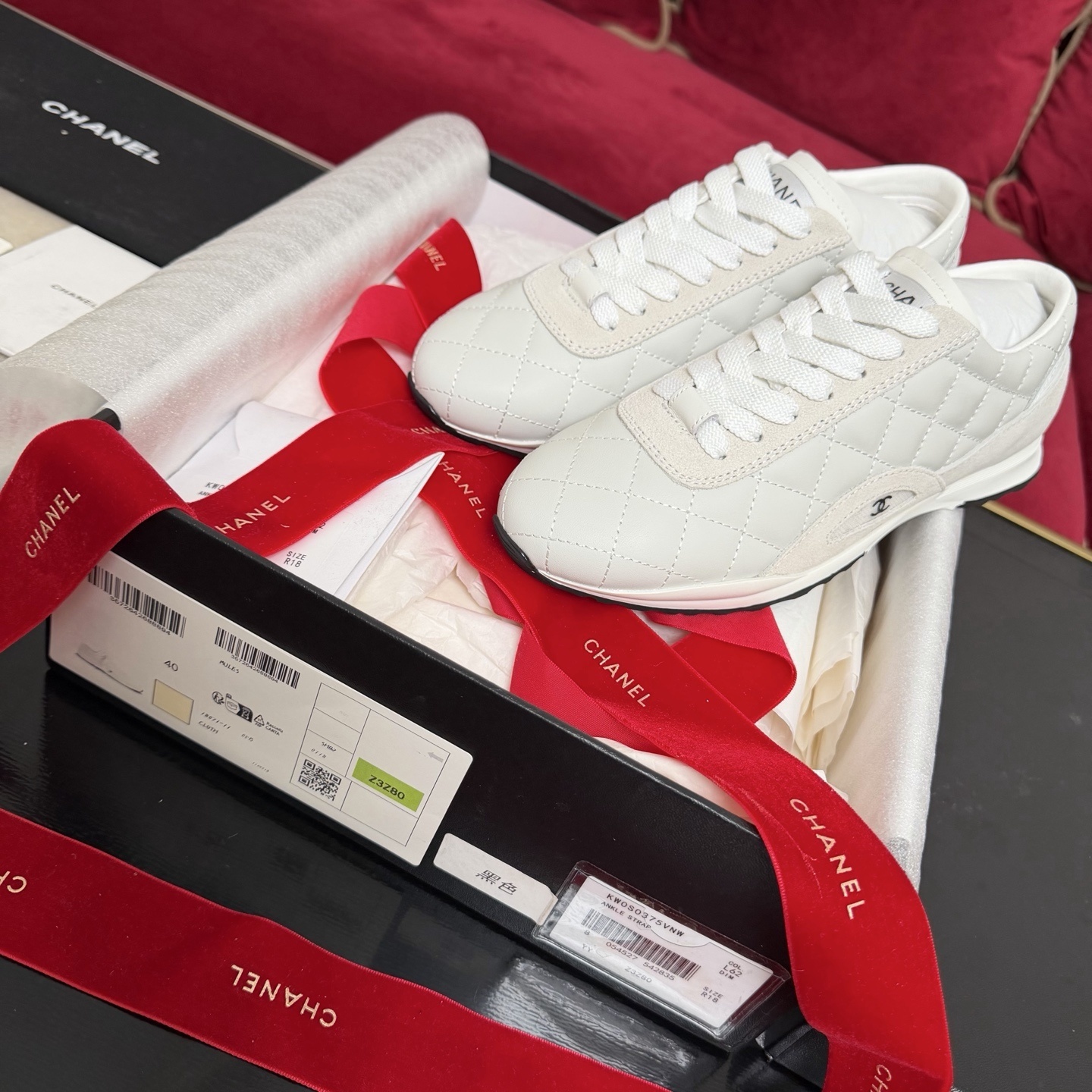 A pair of luxury white Chanel quilted leather sneakers displayed with original black box and decorative red ribbons. - Hình 2