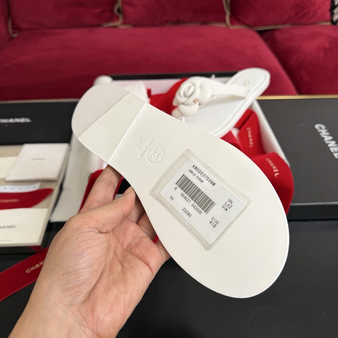 A pair of luxury white Chanel thong sandals featuring a large camellia flower with a gold CC logo on the strap, presented in a black box with red ribbons. - Hình 9