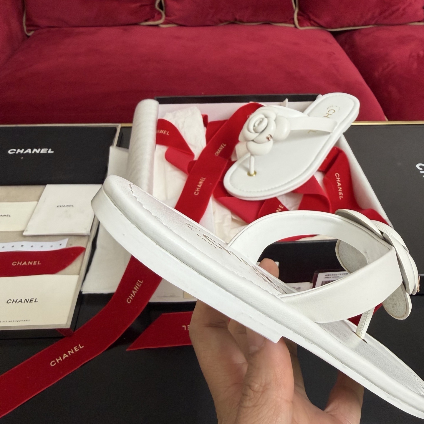 A pair of luxury white Chanel thong sandals featuring a large camellia flower with a gold CC logo on the strap, presented in a black box with red ribbons. - Hình 8