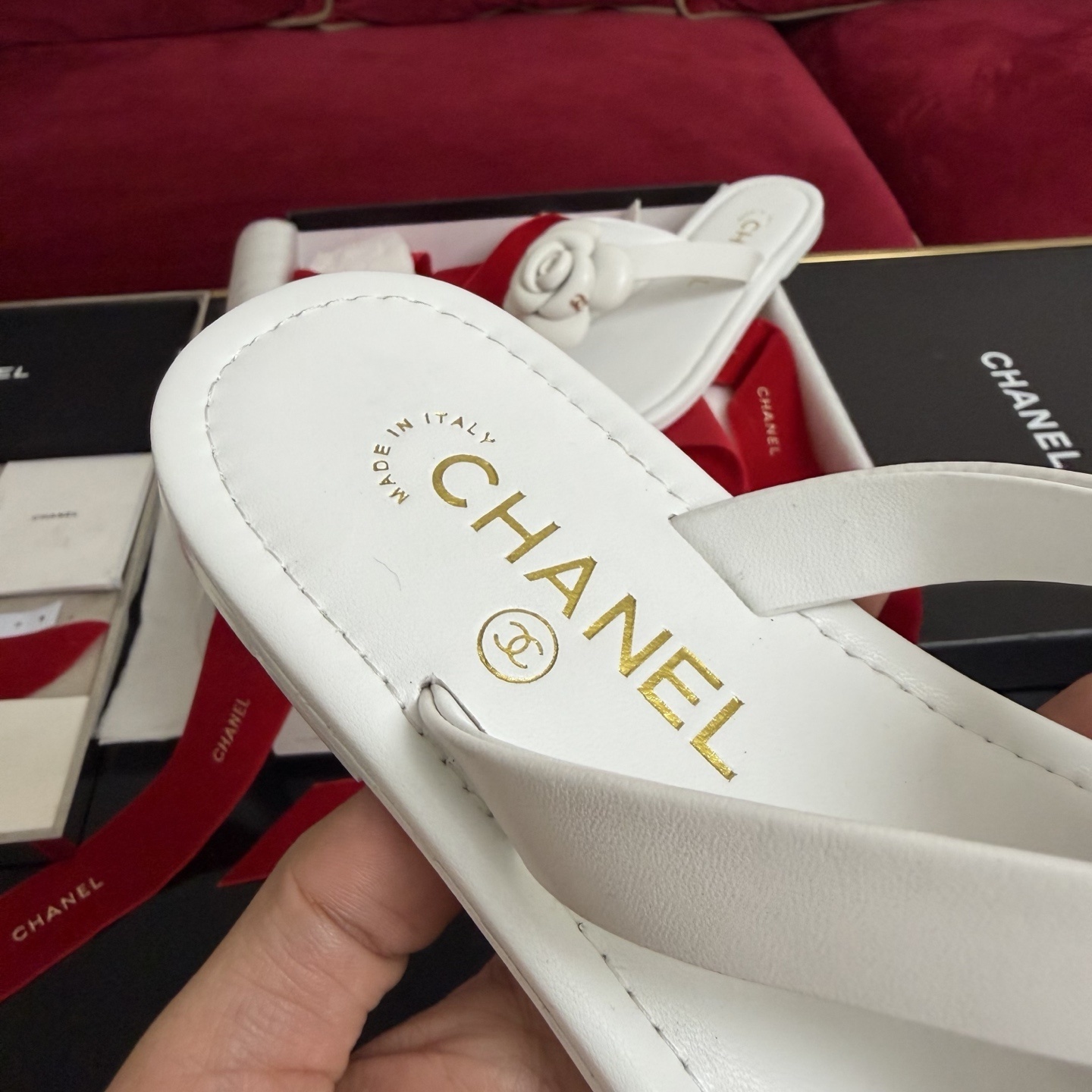 A pair of luxury white Chanel thong sandals featuring a large camellia flower with a gold CC logo on the strap, presented in a black box with red ribbons. - Hình 7