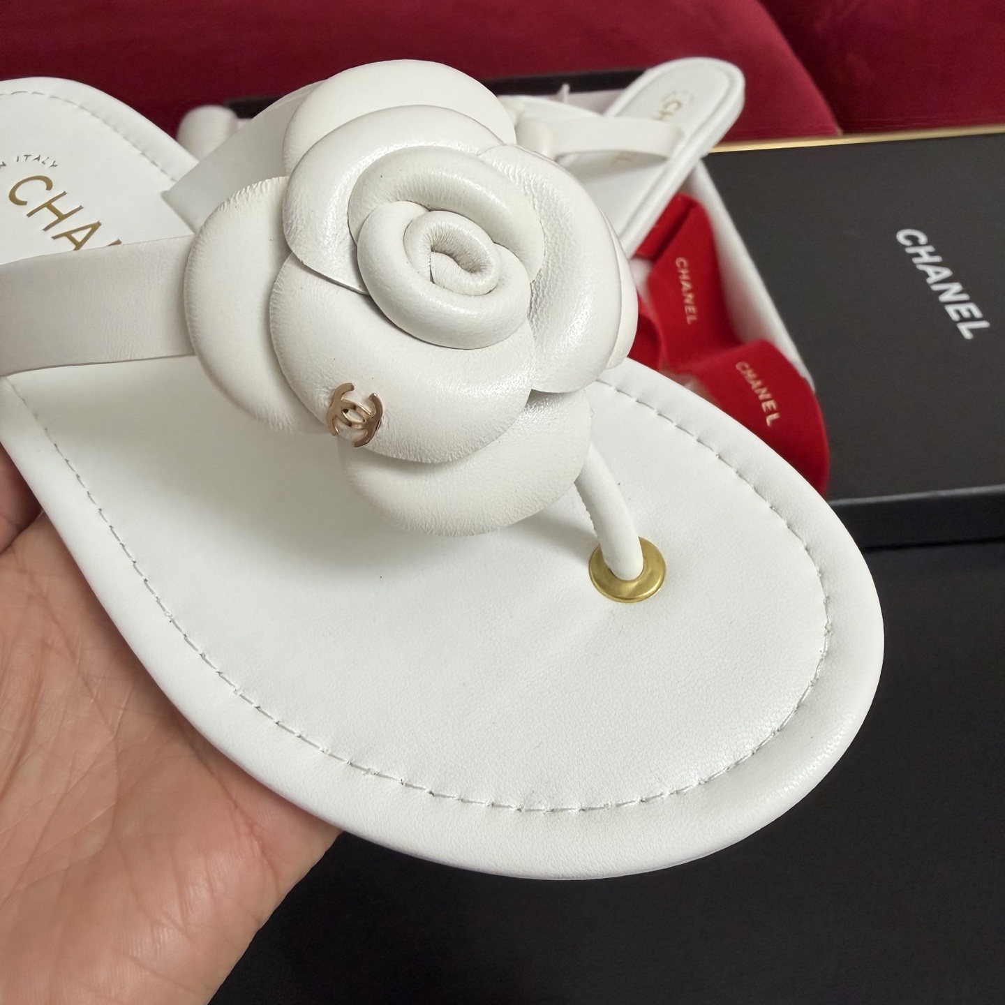 A pair of luxury white Chanel thong sandals featuring a large camellia flower with a gold CC logo on the strap, presented in a black box with red ribbons. - Hình 6