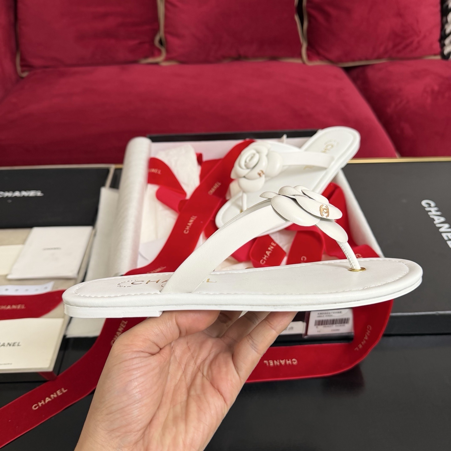 A pair of luxury white Chanel thong sandals featuring a large camellia flower with a gold CC logo on the strap, presented in a black box with red ribbons. - Hình 5