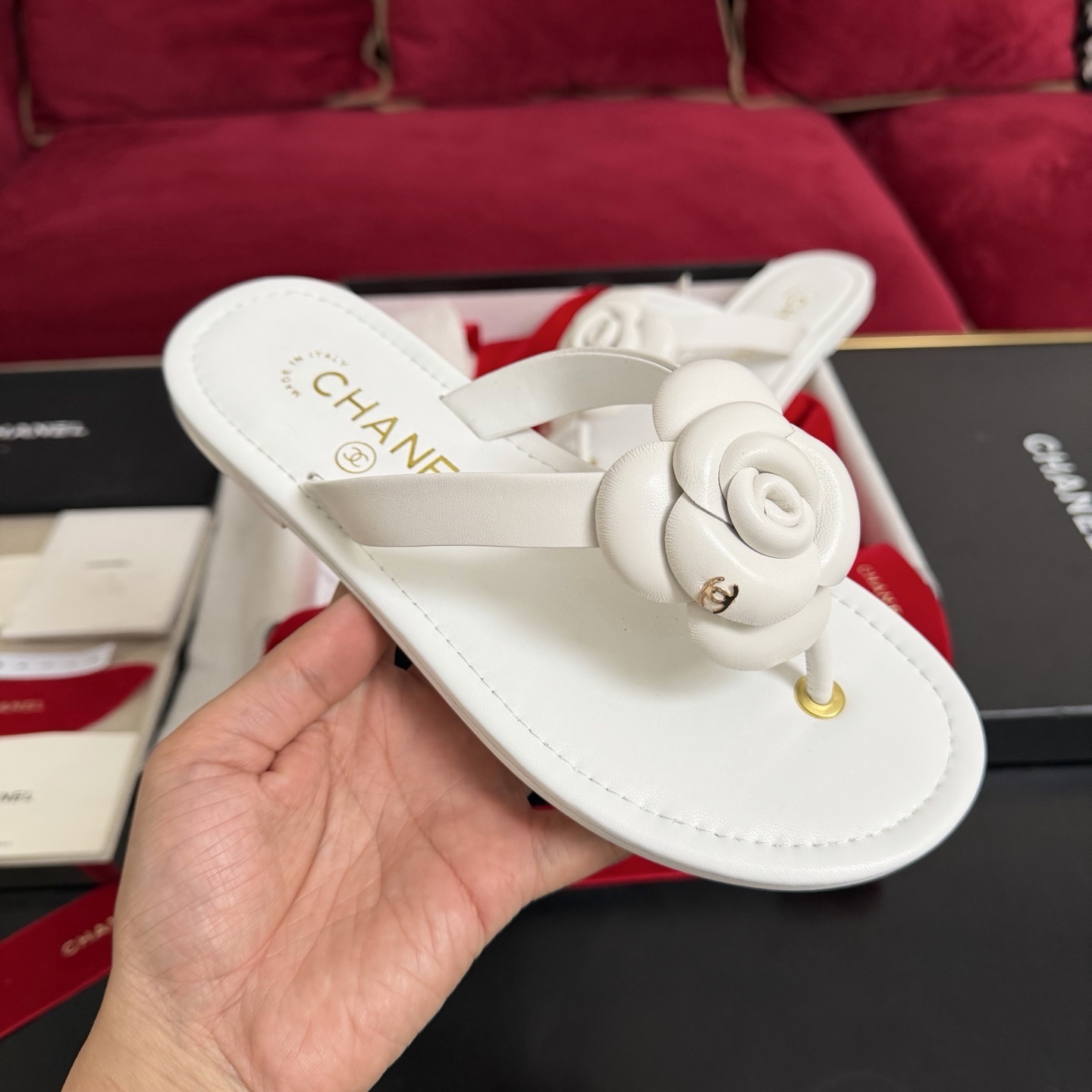 A pair of luxury white Chanel thong sandals featuring a large camellia flower with a gold CC logo on the strap, presented in a black box with red ribbons. - Hình 4